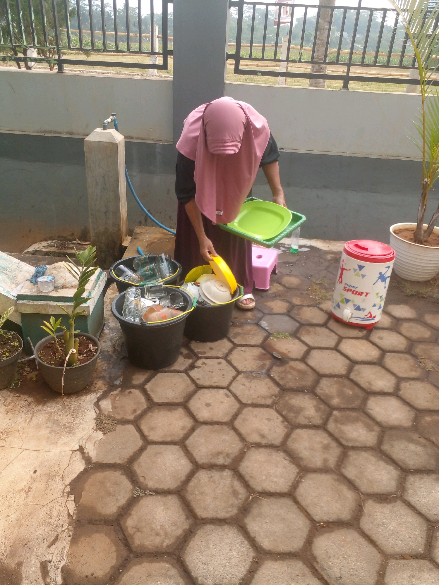 A person wearing a pink hijab and maroon dress is washing dishes in Banyuputih, Indonesia. The person is standing outdoors on a hexagonal paver patio. To the person's left, there are three black buckets overflowing with dishes. A green plastic tray filled with dishes is held by the person, and a yellow plastic bowl is also visible. A pink stool is placed behind the person. To the right, a red and white cooler with "Super Sport" written on it is standing. A potted plant with green leaves is also to the right of the person. In the background, there is a concrete wall, a metal fence, and vegetation.