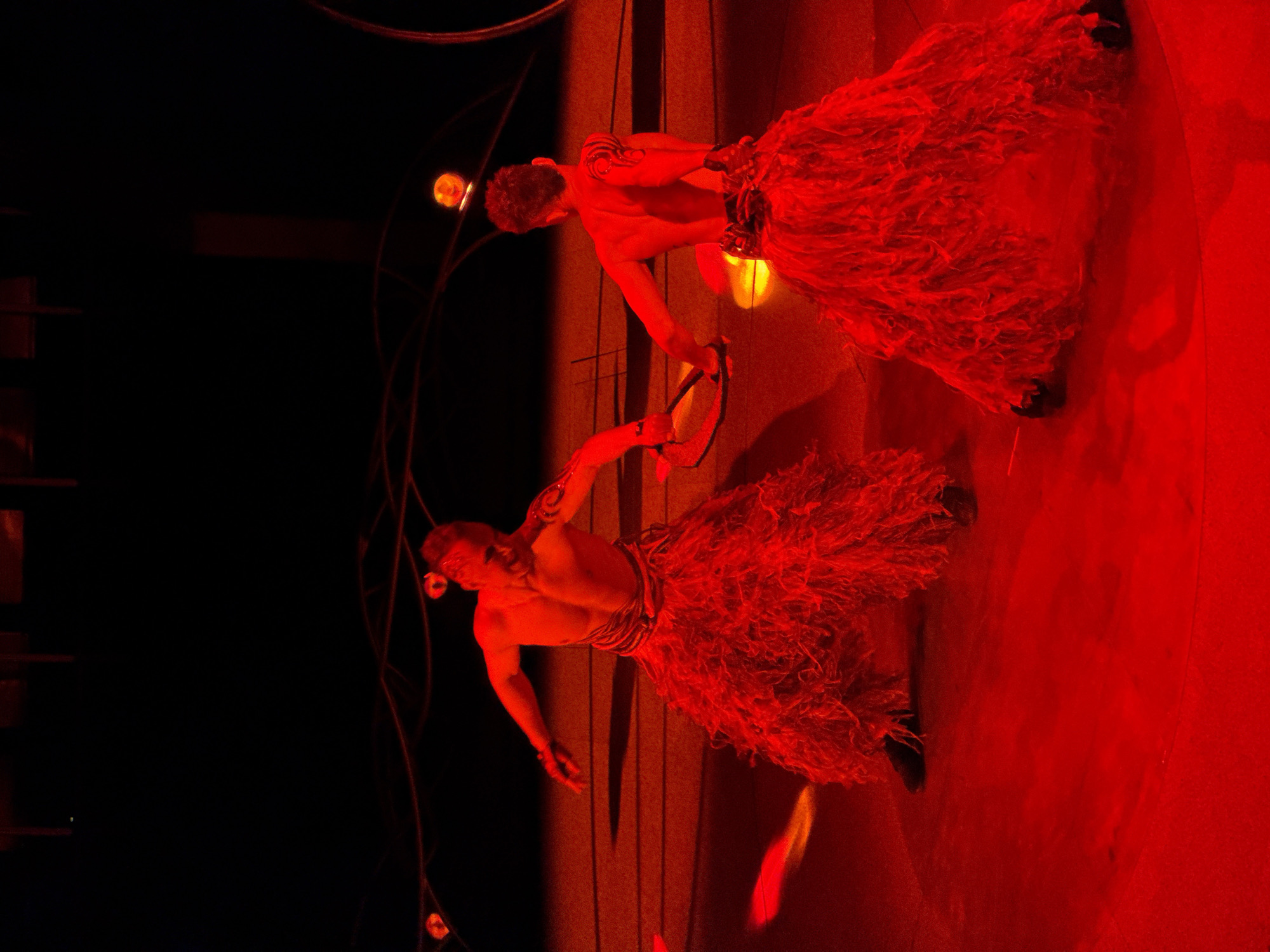 The image shows two shirtless men in red-lit stage setting, possibly in Yerevan, Armenia. They are wearing textured skirts or kilts, and one holds a hoop-like object. The background is a mix of deep black and red-lit areas, with stage elements and lighting visible.