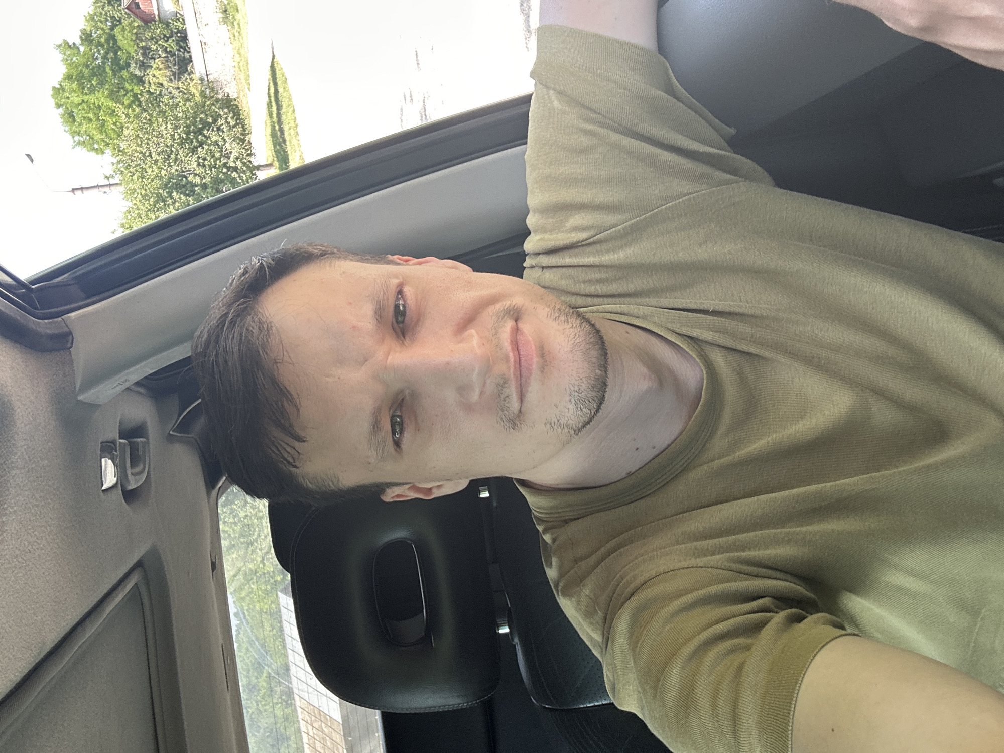 A Caucasian male with short brown hair and stubble is shown from the chest up, sitting in the driver's seat of a car. He is wearing a light green t-shirt. The interior of the car is visible, including a black headrest and part of the dashboard. Through the open car window on the right, a tree-lined street is visible with buildings in the background. The scene appears to be in Makiv, Ukraine, on a sunny day.