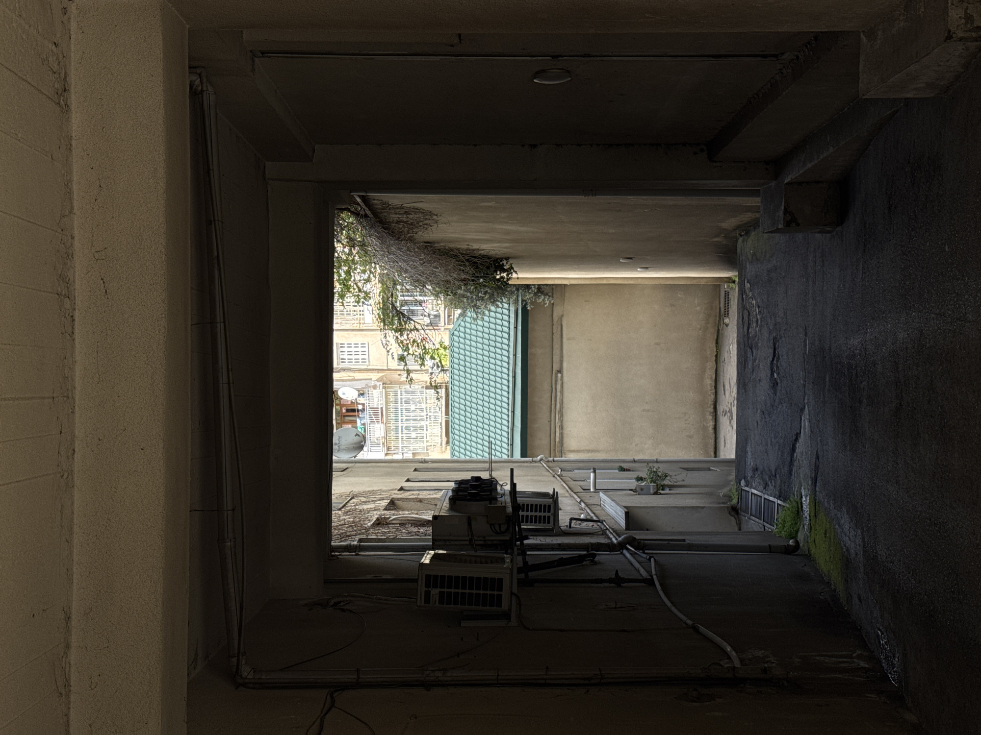 An upward-angled view from a dark, concrete passageway in Tbilisi, Georgia, reveals a courtyard. The passageway is framed by rough, light-colored concrete walls on the left and a dark, possibly wet concrete wall on the right. Overhead, the ceiling is dark and appears to be made of concrete. The courtyard is visible through the opening, showing apartment buildings with windows, a satellite dish, and a green corrugated awning. Several air conditioning units are mounted on the wall, and pipes run along the courtyard floor. Some vegetation, including a plant and moss, are visible in the courtyard area.