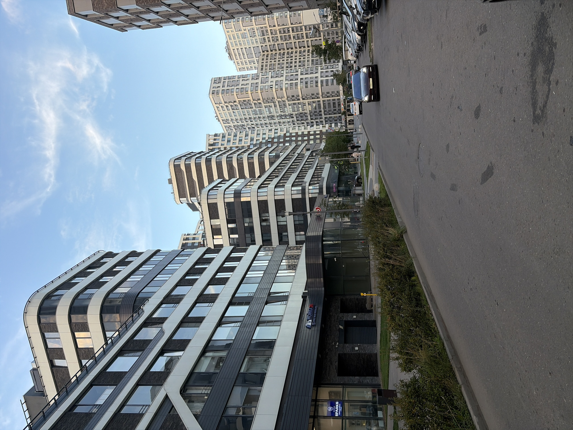 A street scene in Moscow, Russia, captures a row of modern, multi-story residential buildings with distinctive curved facades and dark-colored brickwork interspersed with white horizontal banding. The buildings are tall and contemporary, some featuring balconies. In the foreground, a paved road runs alongside a sidewalk bordered by manicured green bushes and grass. Several parked cars, including a dark sedan in the street and a row of vehicles on the right side, are visible. A few pedestrians are seen walking along the sidewalk in the distance. The sky is partly cloudy, with wispy white clouds against a pale blue backdrop, suggesting it is either early morning or late afternoon. One building has a sign that reads "МОЙКА 24" with a blue logo and a smaller sign with "SPARKS".