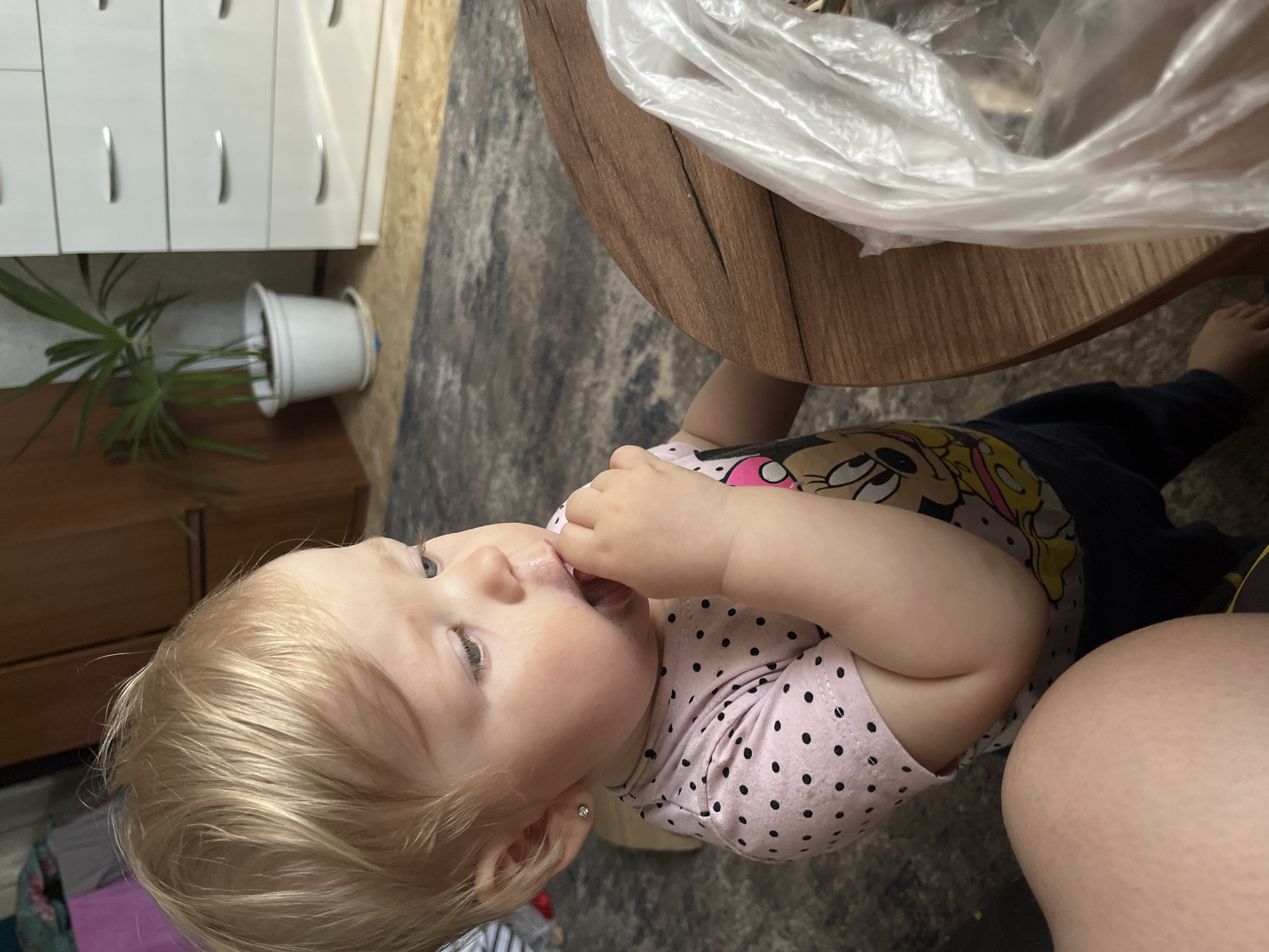 A young Caucasian child with blonde hair and blue eyes is standing and looking down at a round wooden table. The child is wearing a pink shirt with black polka dots and a Minnie Mouse graphic. The child's left hand is in their mouth, and their right hand is on the table. A white plastic bag is on the table next to the child. In the background, there is a white dresser and a potted plant. The floor is covered with a patterned rug. The scene appears to be an indoor setting, possibly a home, in the city of Fokino, Russia.