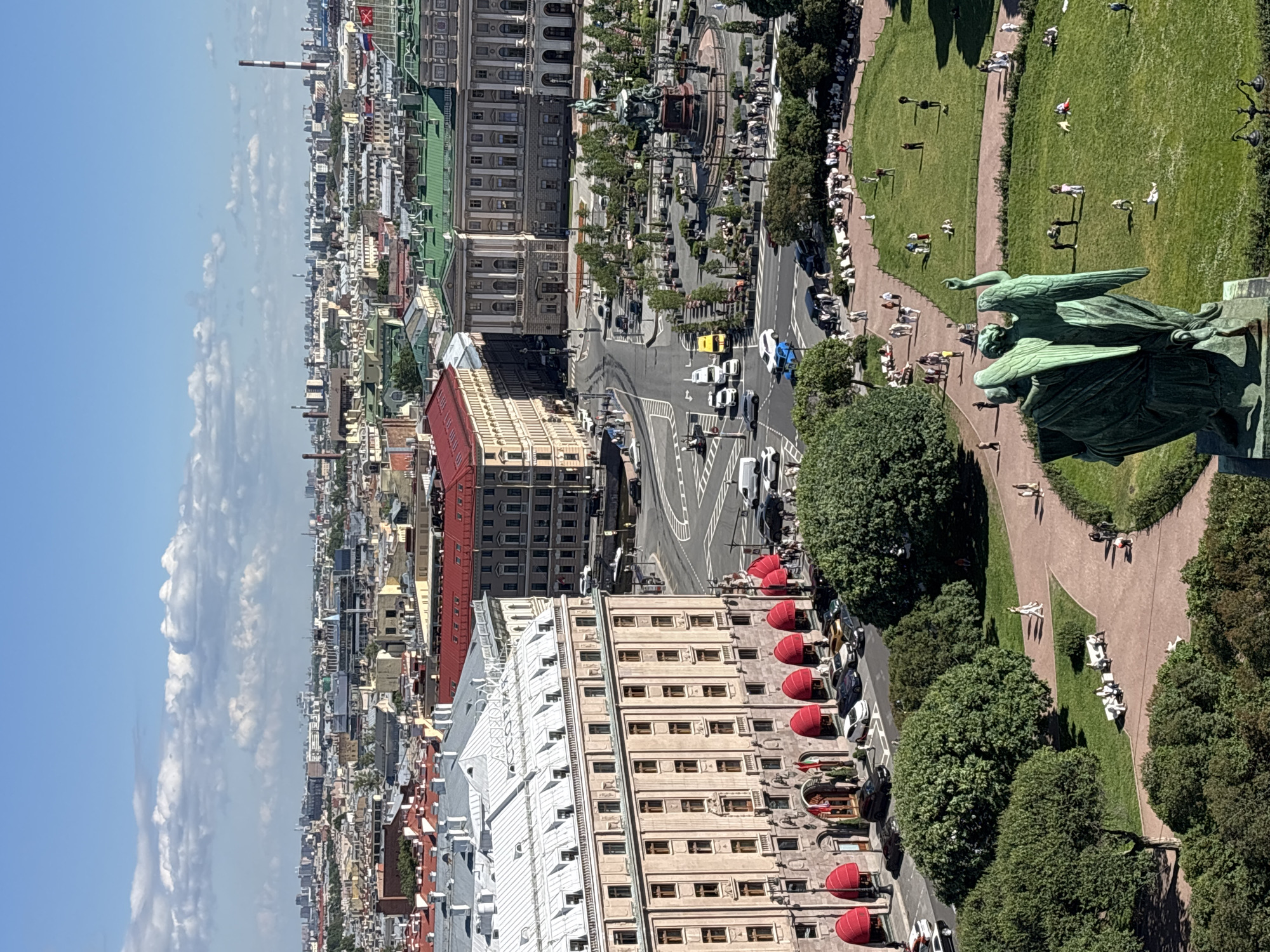 This is a high-angle, vertical aerial photograph of a city on a sunny day, likely in Saint Petersburg, Russia, given the architectural style. The scene captures a sprawling cityscape with a mix of historical and modern buildings, characterized by varied rooflines and facades. In the foreground, a large, green bronze statue of an angel with outstretched wings stands prominently, pointing to the right.
Below the statue, a park with lush green grass and paved pathways is visible, with people scattered throughout, some walking, others resting on benches. Trees line the park's edges and further into the city, creating a blend of urban and natural elements. A road runs alongside the park, with cars and what appears to be a yellow taxi moving along it. The buildings are a mix of colors, including red, white, beige, and green, with some featuring ornate architectural details and awnings. In the distance, a tall red and white chimney stack is a distinct landmark. The sky is a bright blue with scattered white clouds, suggesting a pleasant daytime atmosphere. The overall impression is a vibrant, lively urban environment with areas of public relaxation and architectural grandeur.