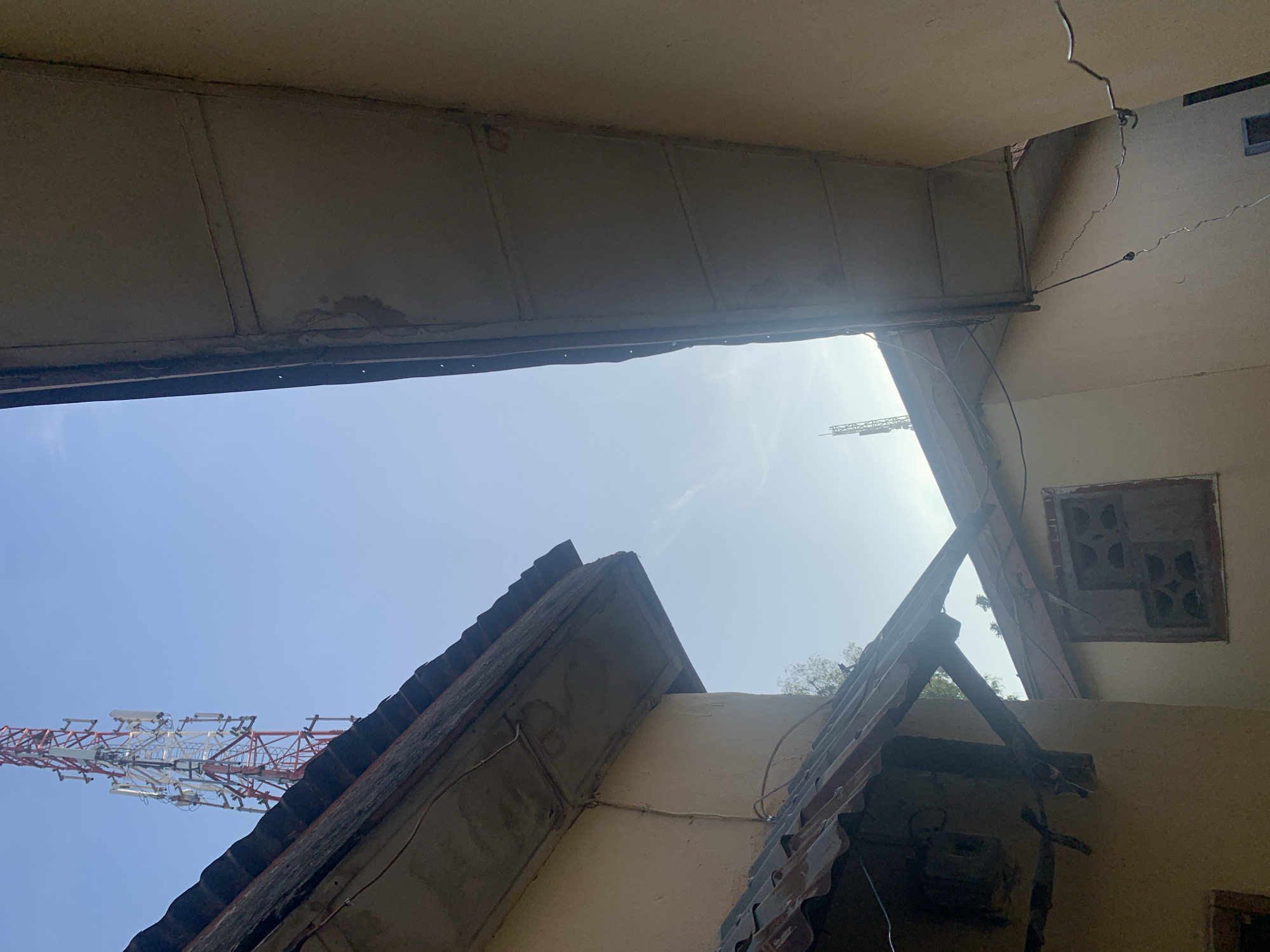 The image captures a narrow alley between two buildings, with a large telecommunications tower visible in the background against a clear blue sky. The buildings appear to be constructed with yellow-colored walls and have corrugated metal roofs that show signs of age and wear. One building on the left has an exposed overhang with rusted metal sheeting. On the right, another building's ceiling, made of what appears to be plasterboard and supporting beams, is visible. Wires are strung between the buildings and along their exteriors. A window with a decorative, possibly wooden, lattice is visible on the left building. The overall impression is one of a densely built urban environment, possibly in a developing area, characterized by functional but weathered infrastructure. The specific location of Rafin Par, Nigeria, suggests a setting within that region.