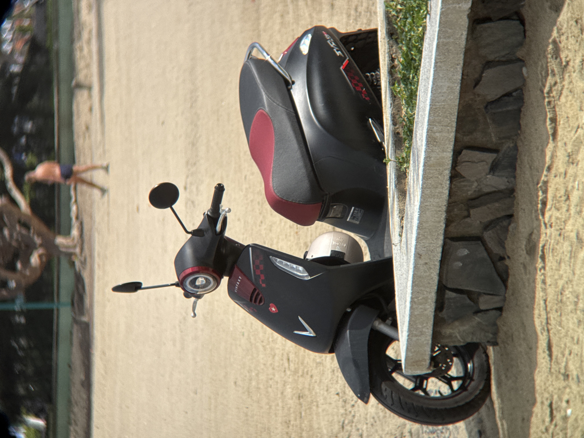The image shows a black VinFast electric scooter parked on a sandy area next to a stone wall. The scooter has a two-tone seat with red accents. A white helmet is attached to the side of the scooter. In the background, a person wearing dark blue shorts walks away from the camera. The environment appears to be a beach or park area, with sand, vegetation, and trees visible. The scooter has a distinctive "V" logo on the front and red branding on the side panel. The scene is brightly lit, suggesting it is daytime. This scene is consistent with a location in Nha Trang, Vietnam, known for its beaches and coastal areas.