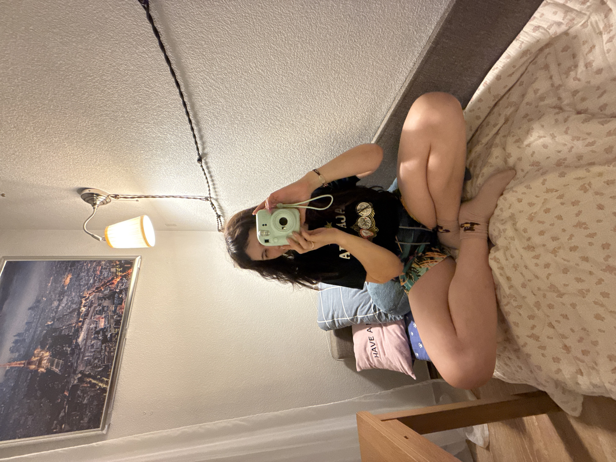 An overhead, eye-level, medium shot shows a young woman with dark, wavy hair sitting cross-legged on a bed. She is wearing a black t-shirt with a graphic on the front and multi-colored shorts. The woman is holding a mint green instant camera up to her face, taking a selfie. She is looking at the camera and smiling. The bed is covered with a white comforter with a floral pattern. To the left of the woman is a pink pillow that says "HAVE A". Behind her are two blue pillows. In the background, a picture of Paris hangs on the wall. A wall lamp with a yellow shade is also visible. The overall scene appears to be an indoor residential setting.