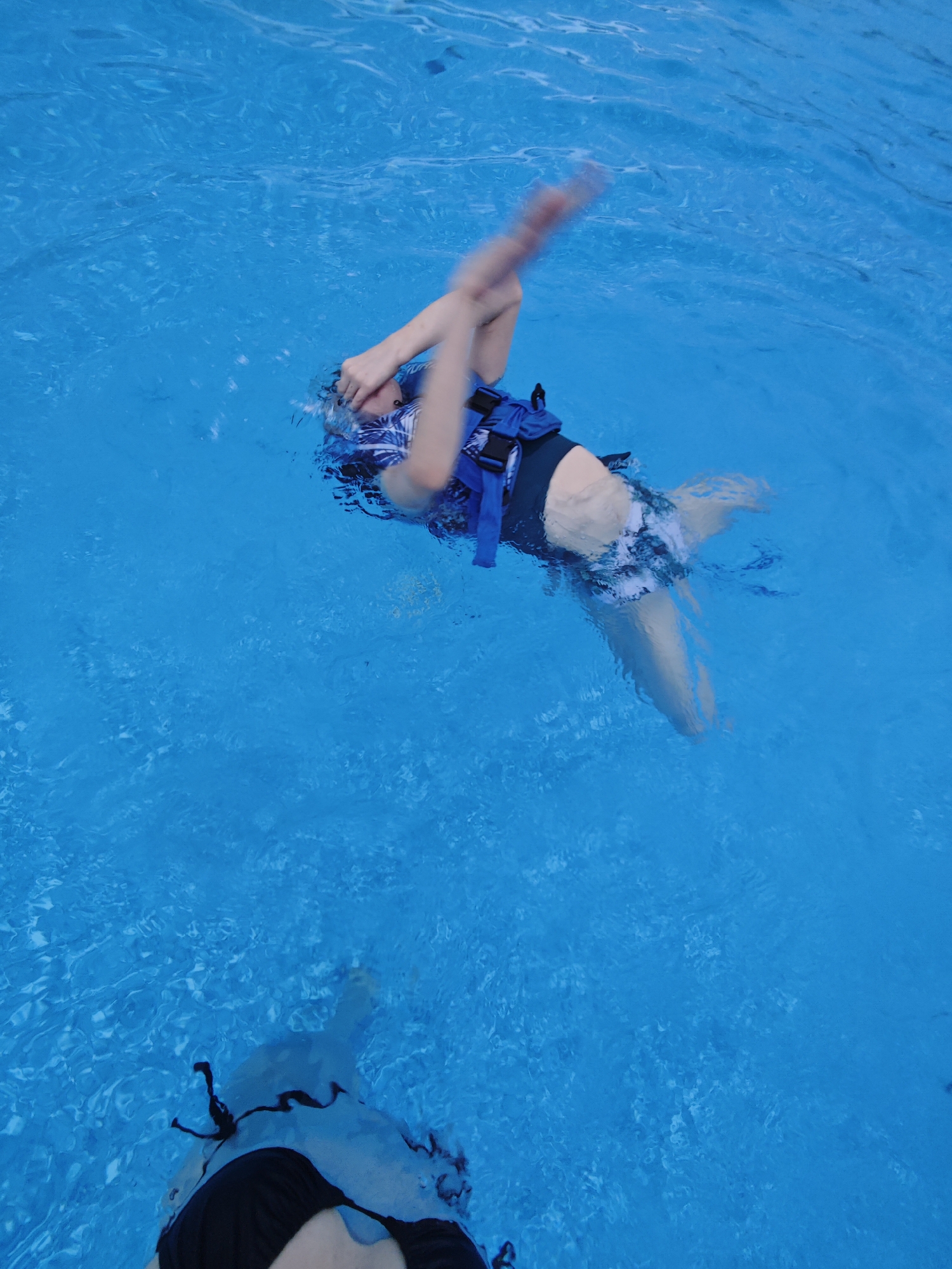 A woman wearing a blue life vest and swimsuit is floating in a swimming pool. Her arms are above her head. At the bottom of the frame, another woman is partially visible, also in the water, wearing a black bikini. The water in the pool is blue and slightly rippled. The location is likely a private or public swimming pool. There is no indication that this photo was taken in Samara, Russia.
