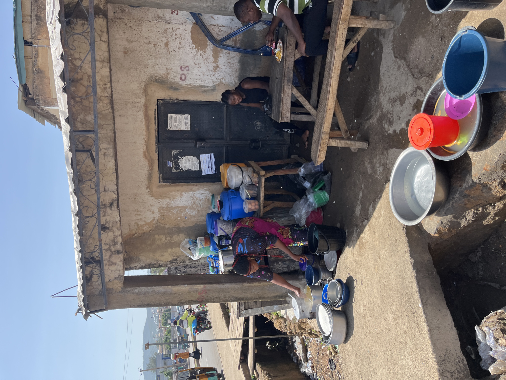 The image captures a vibrant street scene in Rafin Par, Nigeria, under bright daylight with a clear blue sky. A worn, single-story concrete building with a corrugated metal roof dominates the left side of the frame. Its textured, cracked walls feature two dark metal doors. The number "33" is painted in red on the wall to the right of the doors. Papers are affixed to the doors, with one clearly displaying "01510202" and another showing "NPOWER" and a "FG" (Federal Government) logo, possibly indicating a government program or information point.
In front of the building, two individuals are seated at a rustic wooden table. A man, wearing a green and black striped shirt, is seen eating from a plate laden with colorful food items. Beside him, a woman in a black top looks towards the viewer with a slight smile. Near their seating area, a collection of blue plastic containers and other items are stacked against the building wall.
Further to the right and closer to the foreground, another woman, dressed in a vibrant patterned skirt and a dark top, is actively tending to a diverse assortment of pots and buckets. These containers, varying in size and color (silver, blue, red, pink), appear to be filled with water or are being washed/organized.
The immediate ground is concrete, leading to an open drainage ditch on the far right, which contains some debris. In the background, beyond the immediate activity, a street is visible with vehicles including a motorcycle (okada) and a tricycle (keke Napep). Distant buildings and hills are present under the bright, clear sky. The scene depicts everyday activities, possibly related to food preparation, water management, or a small business operation, set against the backdrop of a lively street.