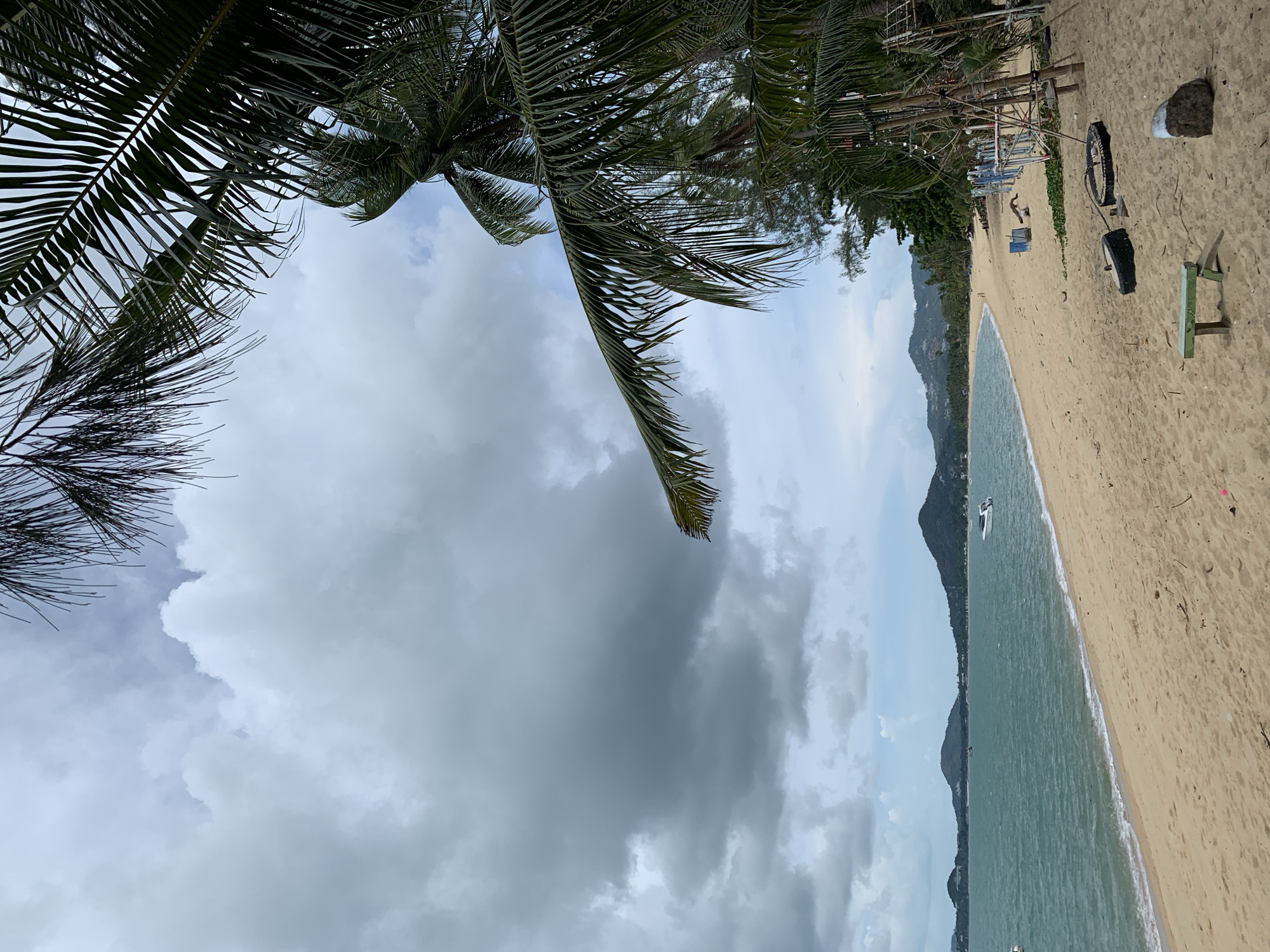 Baan Tai, Thailand. Overcast sky with palm trees in the foreground. A sandy beach stretches along the right side of the image, meeting greenish-blue ocean waters. Mountains are visible across the water. Beachside structures and debris including tires, a bench, and possibly a boat mooring apparatus can be seen in the sand. A single boat is visible on the ocean.