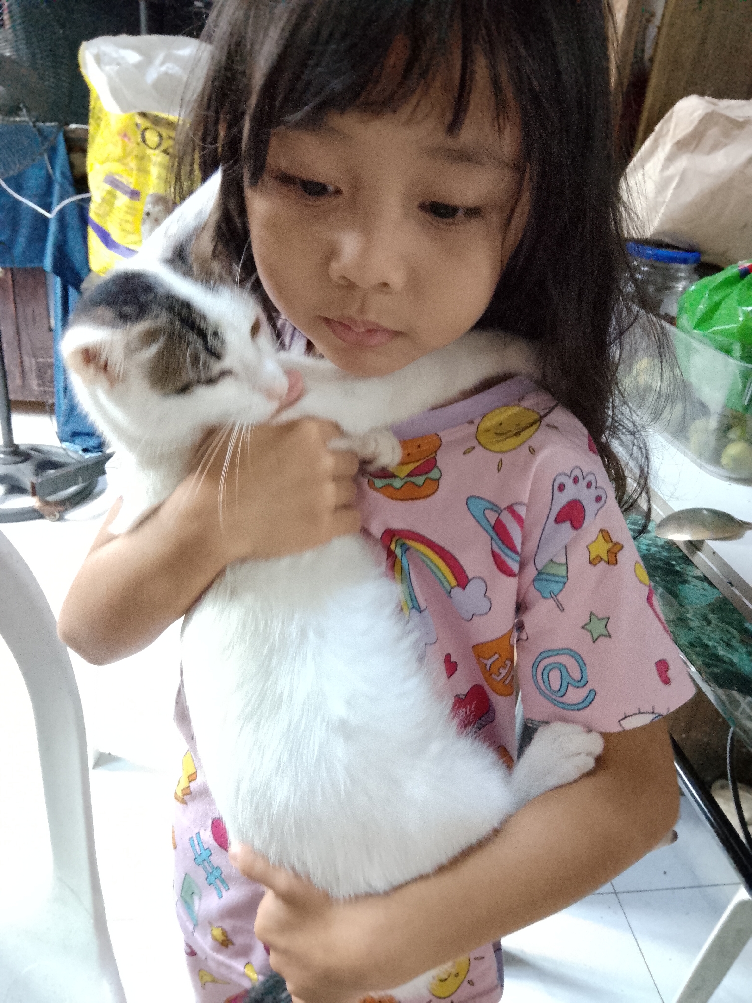 A young child with dark hair is holding a white and brown cat close to their chest. The child is wearing a pink t-shirt with colorful cartoon graphics, including hamburgers, rainbows, planets, and paw prints. The cat is mostly white with brown markings on its head and back, and it appears to be licking the child's cheek. The background is blurred but suggests an indoor setting with some household items and what appears to be a bag of pet food. The scene is likely captured in a residential area within Davao City, Philippines, given the context.