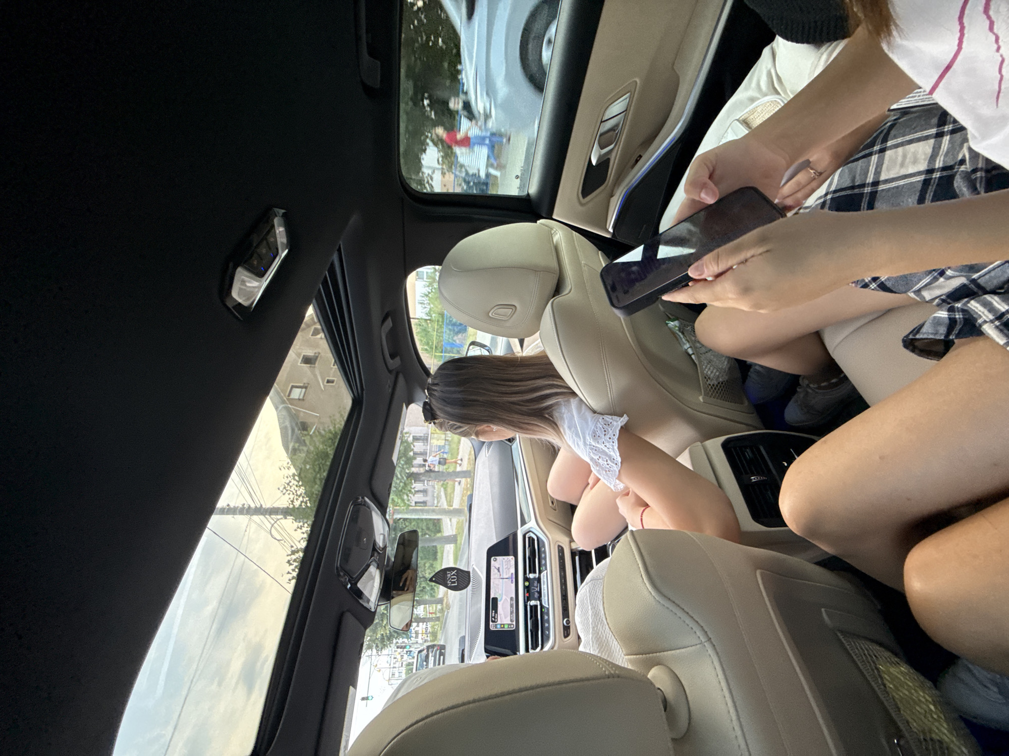 An overhead, wide-angle view captures the interior of a moving vehicle, likely a modern sedan, from the perspective of a rear passenger. The car has a panoramic sunroof, partially open, revealing a hazy sky with clouds and overhead utility lines. The focus is primarily on the rear passenger seats, occupied by three individuals.
In the foreground, two people are visible from the waist down. The person on the left has their legs extended, wearing shorts and a dark plaid skirt. Their arms are not clearly visible. The person on the right, in the center of the frame, is holding a black smartphone in their right hand and is looking down at it. Their left arm is bent, resting on their lap. They are wearing a white top with a pink stripe and a dark plaid skirt.
In the middle ground, a woman with shoulder-length blonde hair is seated in the passenger seat behind the driver. She is wearing a white, short-sleeved top with lace detailing around the neckline. Her arms are bent, and she appears to be looking towards the front of the car or engaging with someone in the front. A black "SPORT LUX" car air freshener hangs from the rearview mirror. The car's dashboard and infotainment screen are visible in the distance, displaying a GPS navigation interface.
In the background, through the car's windows, a street scene unfolds. Buildings with tiled roofs line the street, and trees with green foliage are visible. Utility poles and wires crisscross the sky. Pedestrians are seen walking on the sidewalk, and another vehicle is in motion on the road. The environment suggests an urban or suburban setting during daylight. The image does not provide specific indicators to pinpoint the exact location within Onești, Romania, but the presence of typical street infrastructure and architecture is consistent with such a setting.