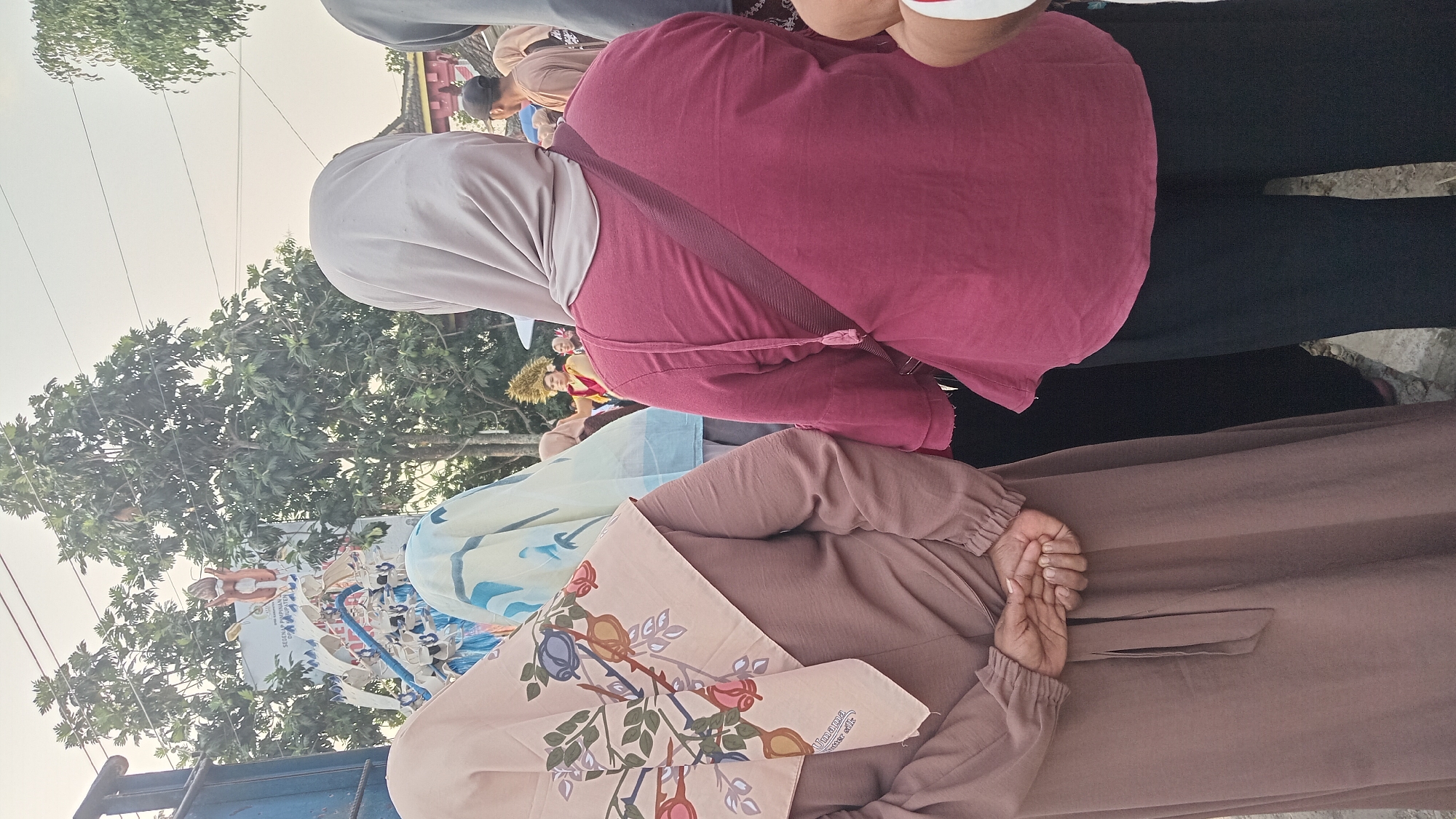 In Kedungwaru, Indonesia, the image captures a street scene with several people. In the foreground, two individuals are partially visible from behind. The person on the left wears a light brown hijab with a floral pattern featuring roses and leaves, and a matching light brown dress. Their hands are clasped behind their back. To their right, another person wears a beige hijab and a maroon top with a dark maroon strap draped diagonally across their back, paired with black pants.
Behind these individuals, other people are seen, some wearing hijabs, and one person in the background wears a distinctive headdress with gold accents. A partially obscured vehicle, possibly a truck, is on the far left. In the background, trees with dense foliage are visible against a bright sky. The ground appears to be a rough, paved surface. The overall impression is of a gathering or procession in an outdoor urban or semi-urban setting.