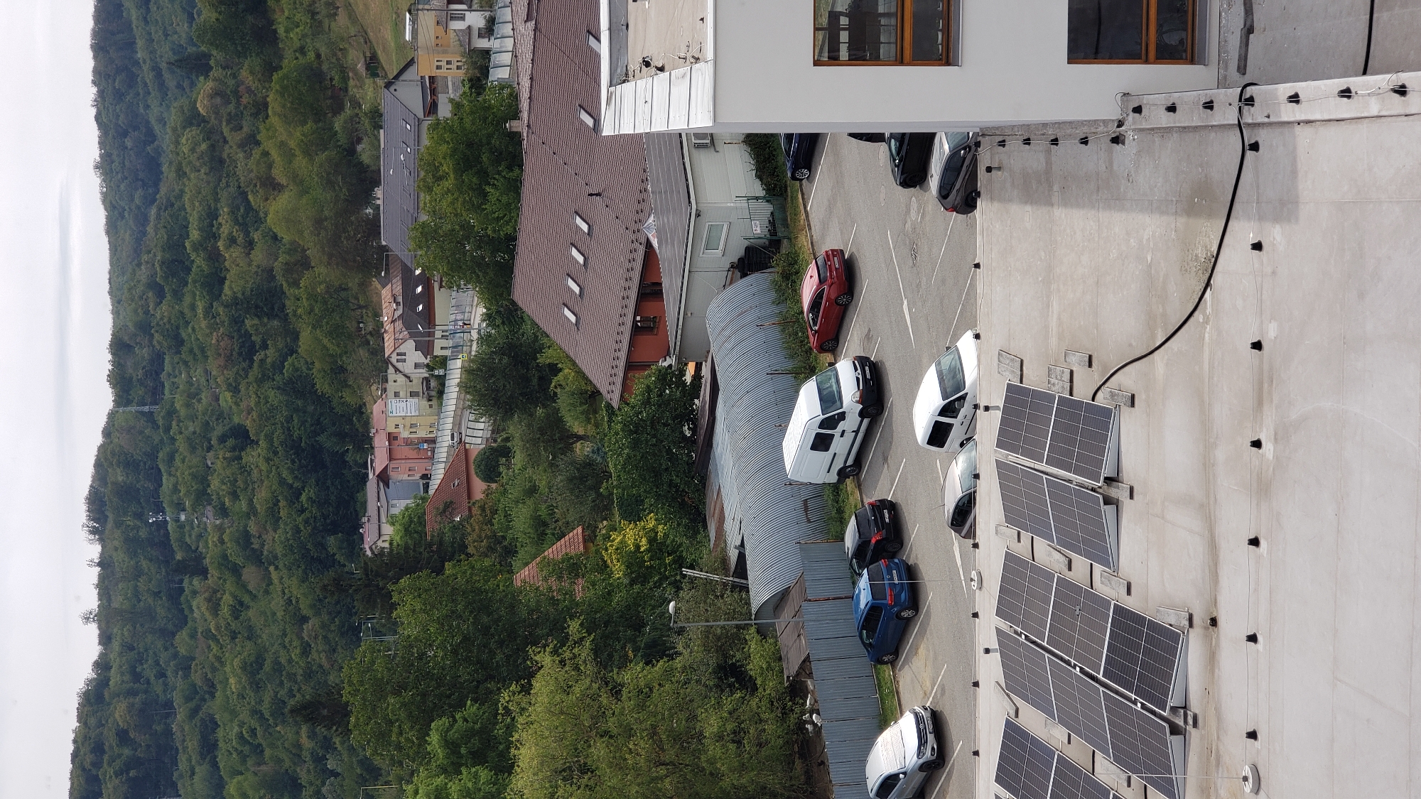 An aerial view captures a parking lot filled with vehicles, situated in front of a cluster of buildings and a forested hillside. The scene appears to be in the city of Prešov, Slovakia, possibly in a suburban or industrial area. In the foreground, a portion of a building's rooftop is visible, featuring a bank of solar panels. The parking lot contains several cars, including silver, blue, black, and red sedans, along with two white vans and a darker colored SUV. Adjacent to the parking lot is a grey, corrugated metal structure, possibly a warehouse or storage facility. Behind this, a residential area with houses of varying colors and architectural styles is visible, with a dense green forest covering the rolling hills in the background. The sky is overcast, suggesting a cloudy day.