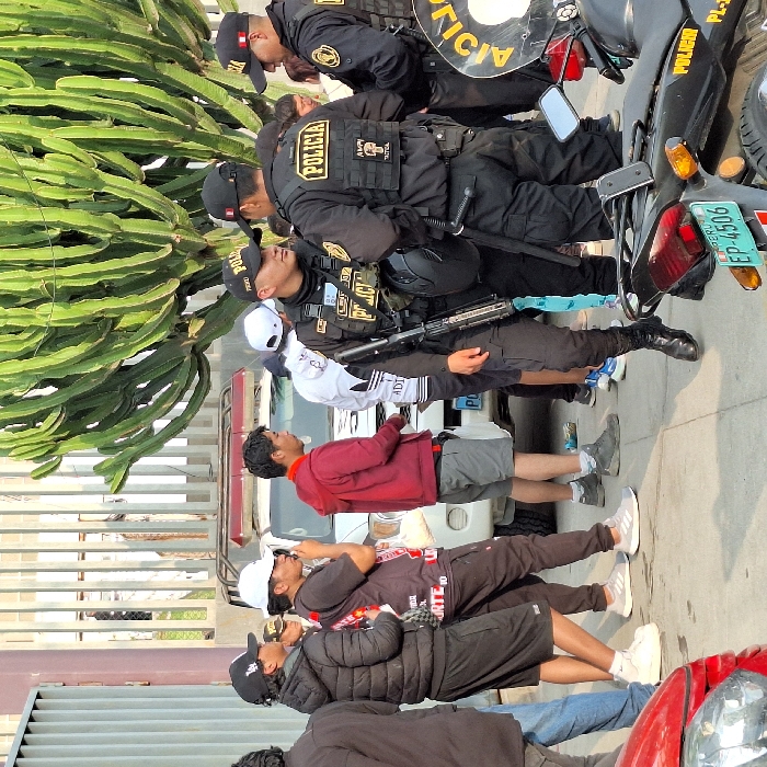 A group of people stand on a street next to several Peruvian police officers in uniform, some with motorcycles. The officers wear tactical vests with "POLICIA" printed on them and helmets. One officer holds a rifle. Civilians stand behind a metal fence, some wearing hoodies and sneakers. A red vehicle is partially visible on the right. In the background, a large cactus-like plant towers over the scene. The license plate on the motorcycle reads "EP-4506."