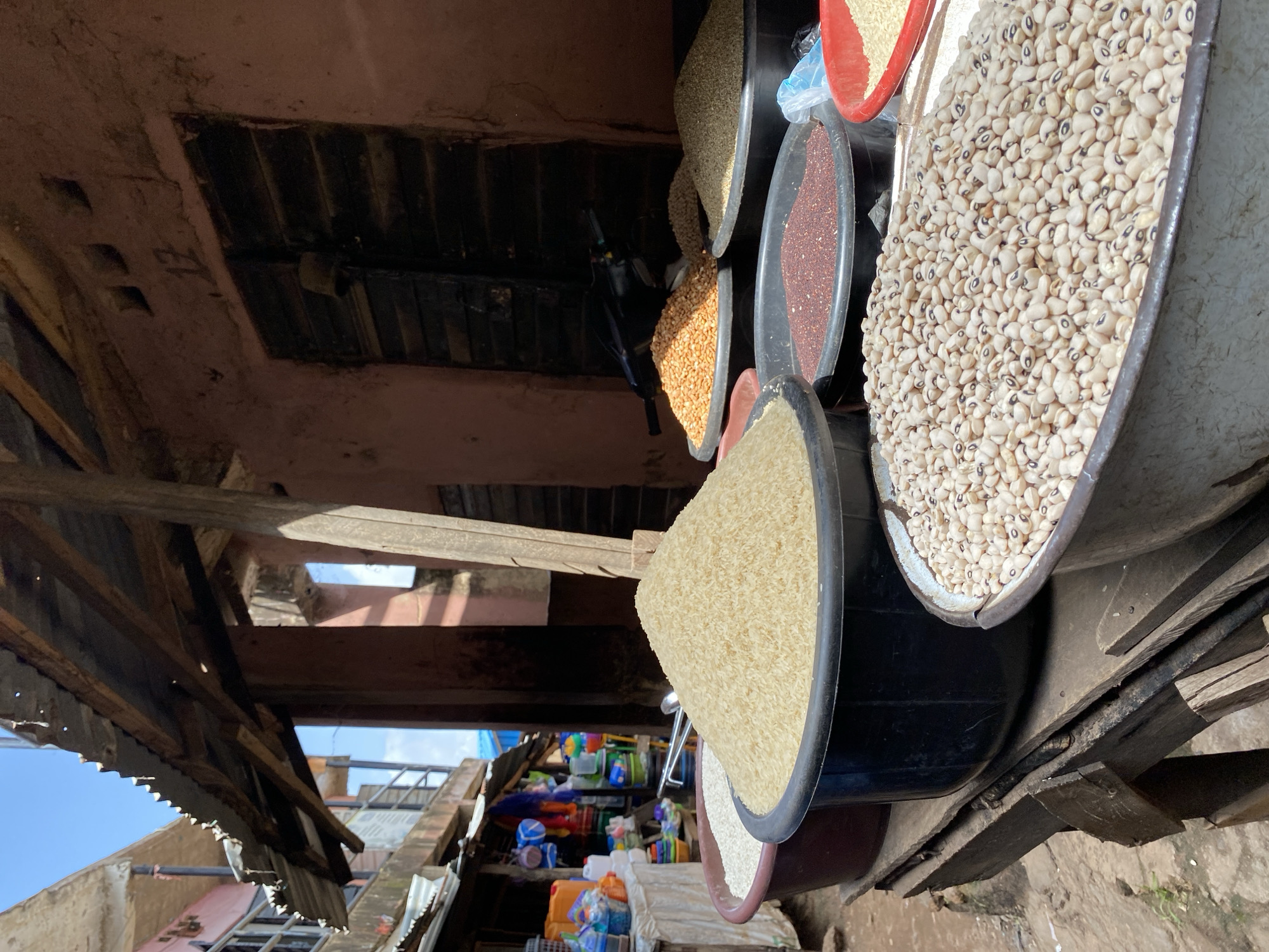 This image captures a vibrant outdoor market scene in Jos, Nigeria. The focus is on a stall displaying a variety of grains and beans in large basins. Prominently featured are overflowing basins of white rice, smaller grains of a reddish-brown hue, and a large quantity of black-eyed peas. Behind these, several other basins hold an assortment of dried goods, including what appears to be maize and lentils. The stall is situated under a makeshift corrugated metal roof supported by wooden beams, suggesting a permanent or semi-permanent market structure. The background reveals the facade of a building painted in earthy tones, with a wooden door and a sign displaying the number "12". To the left, a narrow walkway leads further into the market, with glimpses of other stalls and colorful items for sale. The overall impression is one of bustling commerce in a typical Nigerian urban setting, with the sun casting distinct shadows, indicating it is daytime.
