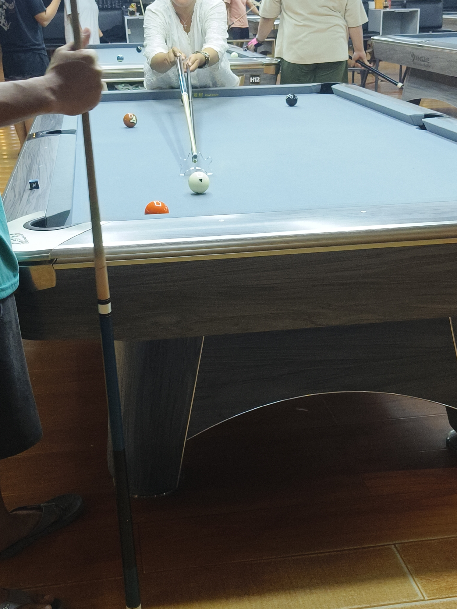 In Kuta Selatan, Indonesia, a billiard table is the central focus of this image. A person's hand, holding a cue stick, is prominently featured in the foreground, indicating an ongoing game. The billiard table itself is light blue with several colored balls scattered across its surface, including a white cue ball and an orange ball. The table has a wooden finish with silver accents along the edge. In the background, other individuals can be seen near the table, with one person appearing to take a shot with a cue stick. The environment appears to be an indoor recreational space, possibly a billiard hall or club, with the floor made of light brown tiles.