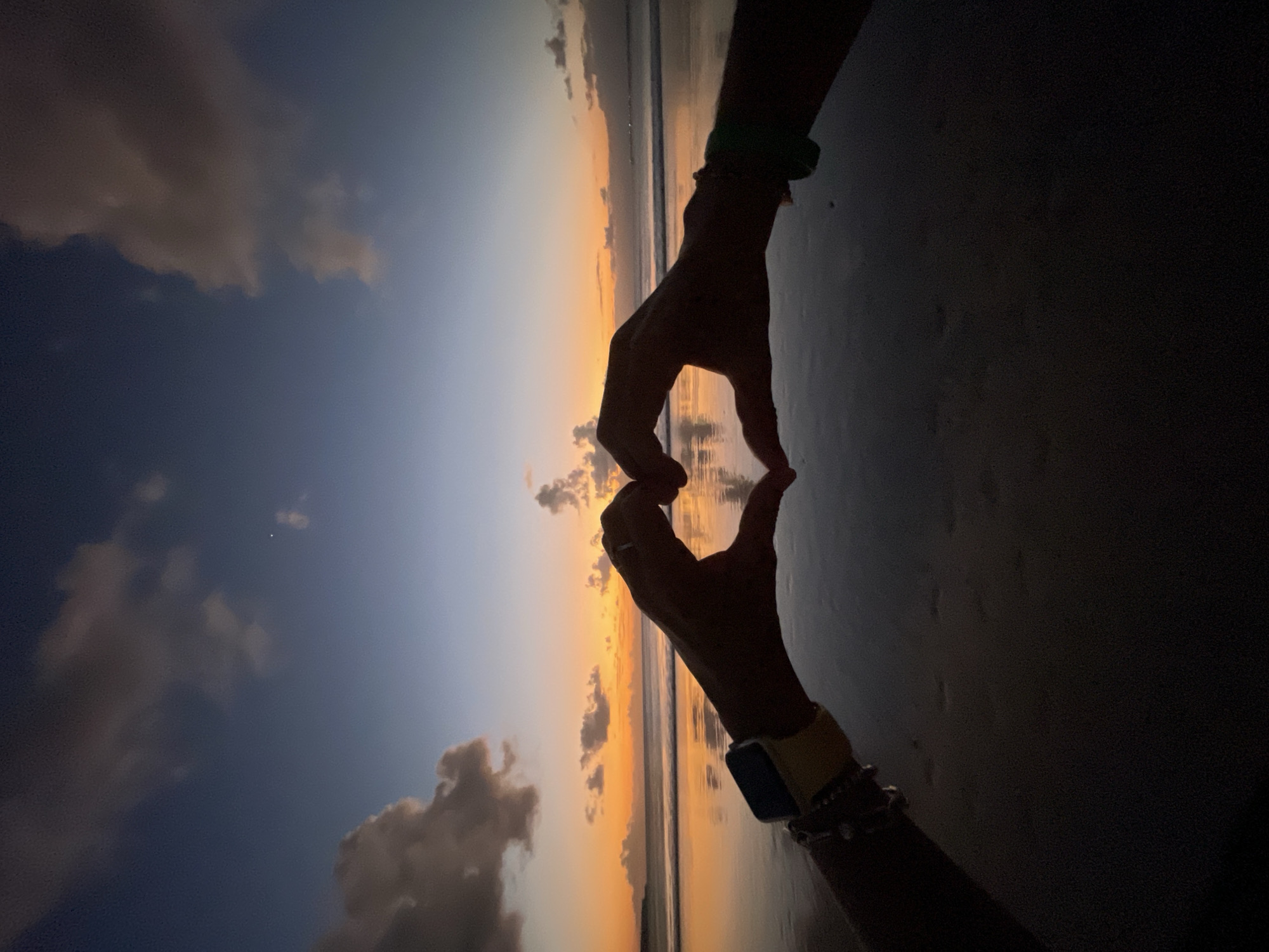 The image depicts a sunset scene at a beach, likely in Kuta Selatan, Indonesia, given the prominent use of warm orange and yellow hues in the sky and their reflection on the wet sand. Two silhouetted hands, belonging to individuals whose faces are not visible, are positioned in the foreground, forming a heart shape. These hands appear to be adorned with bracelets. One wrist is fitted with a yellow-strapped smartwatch. The hands are positioned over the reflective surface of the wet sand, creating a mirrored effect. The ocean is visible in the mid-ground, with gentle waves rolling towards the shore, and the sky above is a gradient of deep blue to bright orange, dotted with scattered clouds. The overall impression is one of romance and tranquility at dusk.