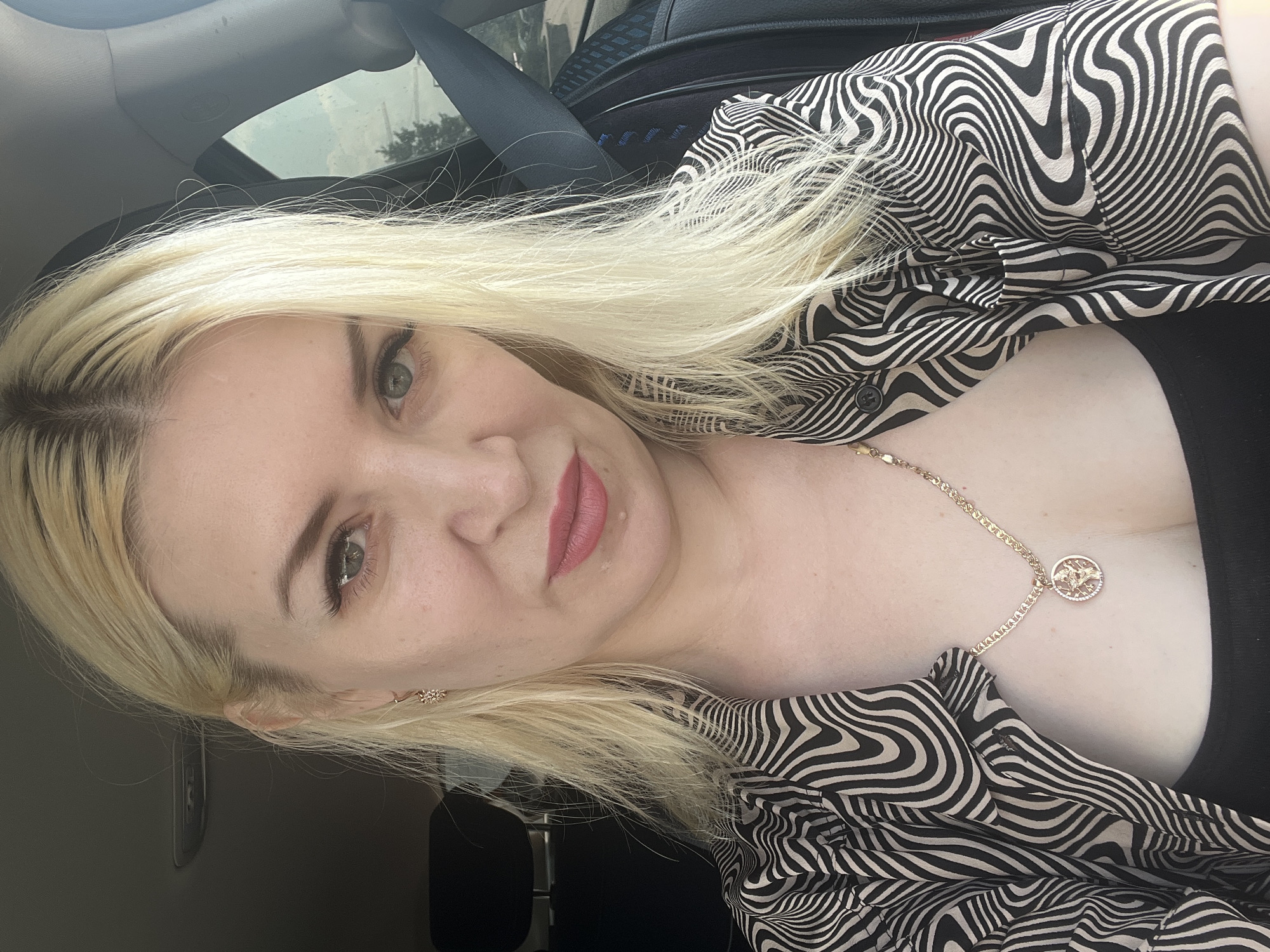 A close-up, selfie-style, medium shot shows a fair-skinned blonde woman in her 30s sitting in the driver's seat of a vehicle. She is wearing a black top underneath an unbuttoned, black and white patterned blouse with a wavy, psychedelic print. A gold chain necklace with a circular pendant rests on her chest. Her blonde hair is partially covering her shoulders. Her eyes are a light green hue, and she has on light makeup, including pink lipstick and mascara. A faint smile is on her face. The car's interior is visible in the background, including a black seat, a seatbelt, and a portion of the passenger seat. The scene appears to be outdoors, with a blurred background suggesting a possibly suburban or highway setting. The image does not provide enough information to ascertain the specific location in Tyumen, Russia, or any specific actions or interactions.