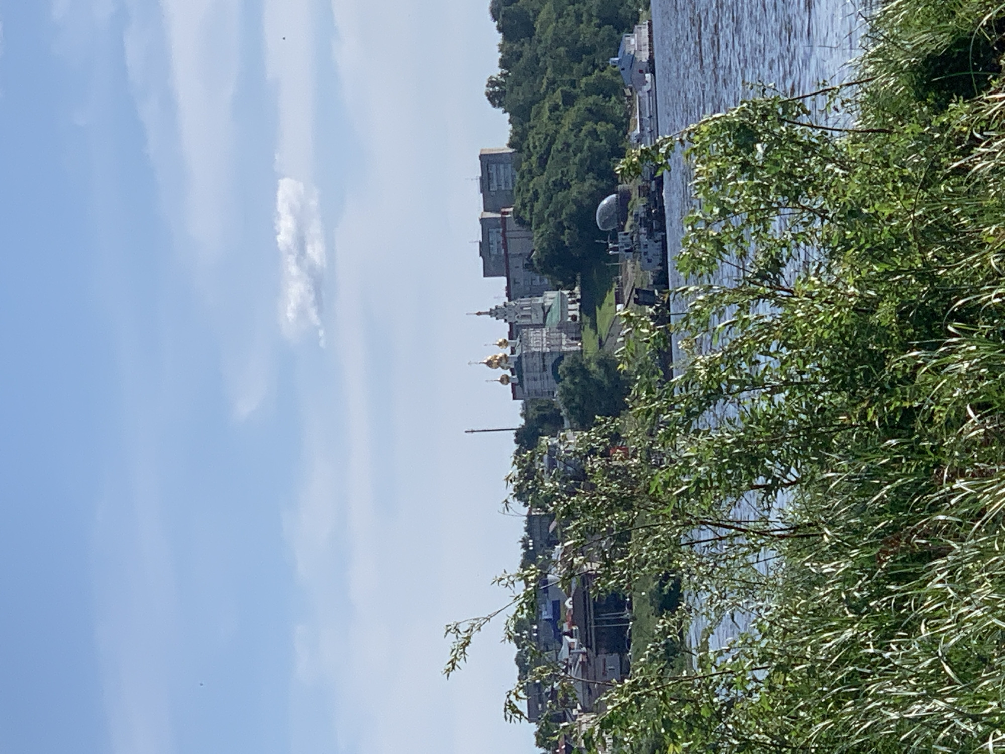 The image depicts a waterside scene in Pavlovo, Russia, likely on a clear, sunny day. The foreground is dominated by lush green foliage, with branches and leaves of trees and bushes creating a natural frame. Beyond the vegetation, a wide body of water, presumably a river, is visible, reflecting the sky and the buildings on the opposite bank.
On the far bank, a cityscape unfolds. Several multi-story buildings, some appearing to be residential or administrative, line the waterfront. Among them, a prominent building with golden domes suggests a church or religious institution. The architecture hints at a historical or culturally significant area. The presence of trees interspersed with the buildings indicates a well-maintained urban environment with green spaces.
Boats are docked or moored along the river, suggesting a functioning port or recreational area. The water's surface is rippled, indicating a gentle breeze. The sky is a clear blue, with a few wispy white clouds. The overall atmosphere is serene and picturesque. The image is oriented vertically, as if captured from a boat or from a low vantage point near the water's edge. No people are clearly discernible in the image, and no text is visible. The scene evokes a sense of tranquility and the beauty of a summer day in a Russian town.
