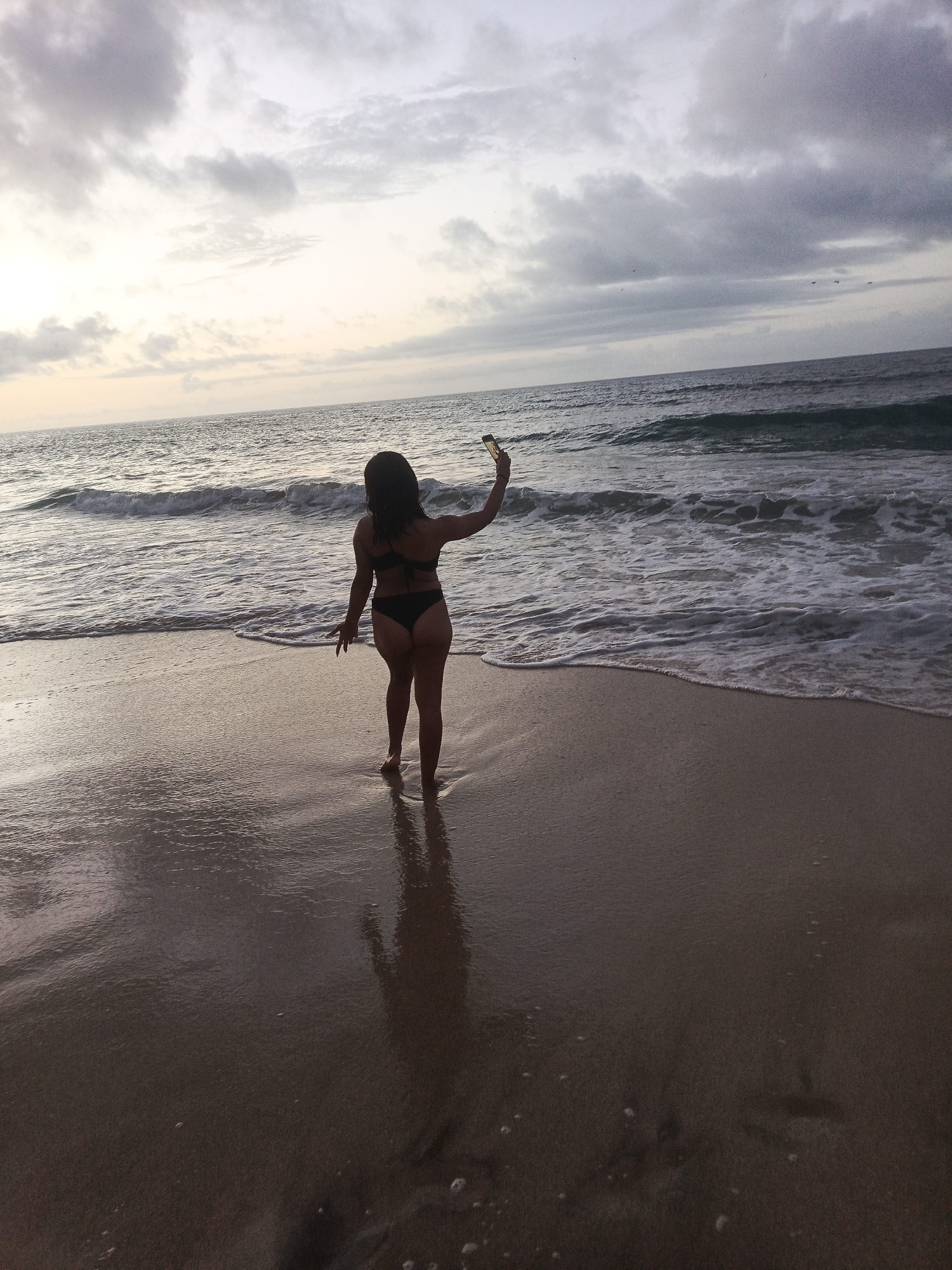 A full shot captures a woman standing on a beach in Pedro González, Venezuela, at dusk. She is facing away from the camera, wearing a black bikini, and holding a smartphone in her right hand as if taking a selfie. The ocean stretches out before her, with waves gently rolling onto the shore. The sky above is a canvas of dramatic clouds, with hints of the setting sun casting a warm glow on the horizon. The wet sand reflects the fading light, creating a mirror-like surface. Footprints are faintly visible on the sand, suggesting previous activity on the beach. The overall atmosphere is serene and contemplative, with the woman appearing to be enjoying a moment of solitude by the sea.