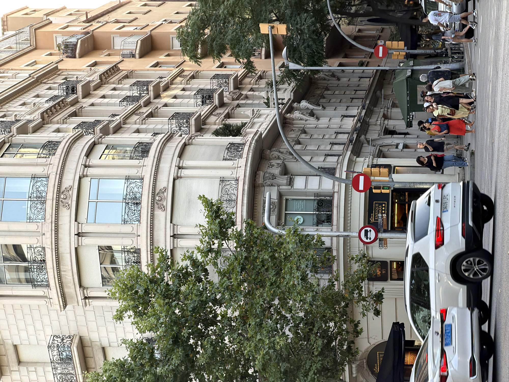 The image shows a street scene in Barcelona, Spain, likely on a warm, overcast day. In the foreground, a white SUV, possibly a Seat Ateca, is driving on the right side of the road. To its left, the rear of another white car is visible. Several people are crossing the street at a pedestrian crossing. One woman in a red dress is prominent in the group. The background features a multi-story building with ornate architecture, including balconies with decorative iron railings and arched windows. A large tree with green foliage partially obscures the building. Traffic signals and street signs are visible. The overall impression is a bustling urban environment.