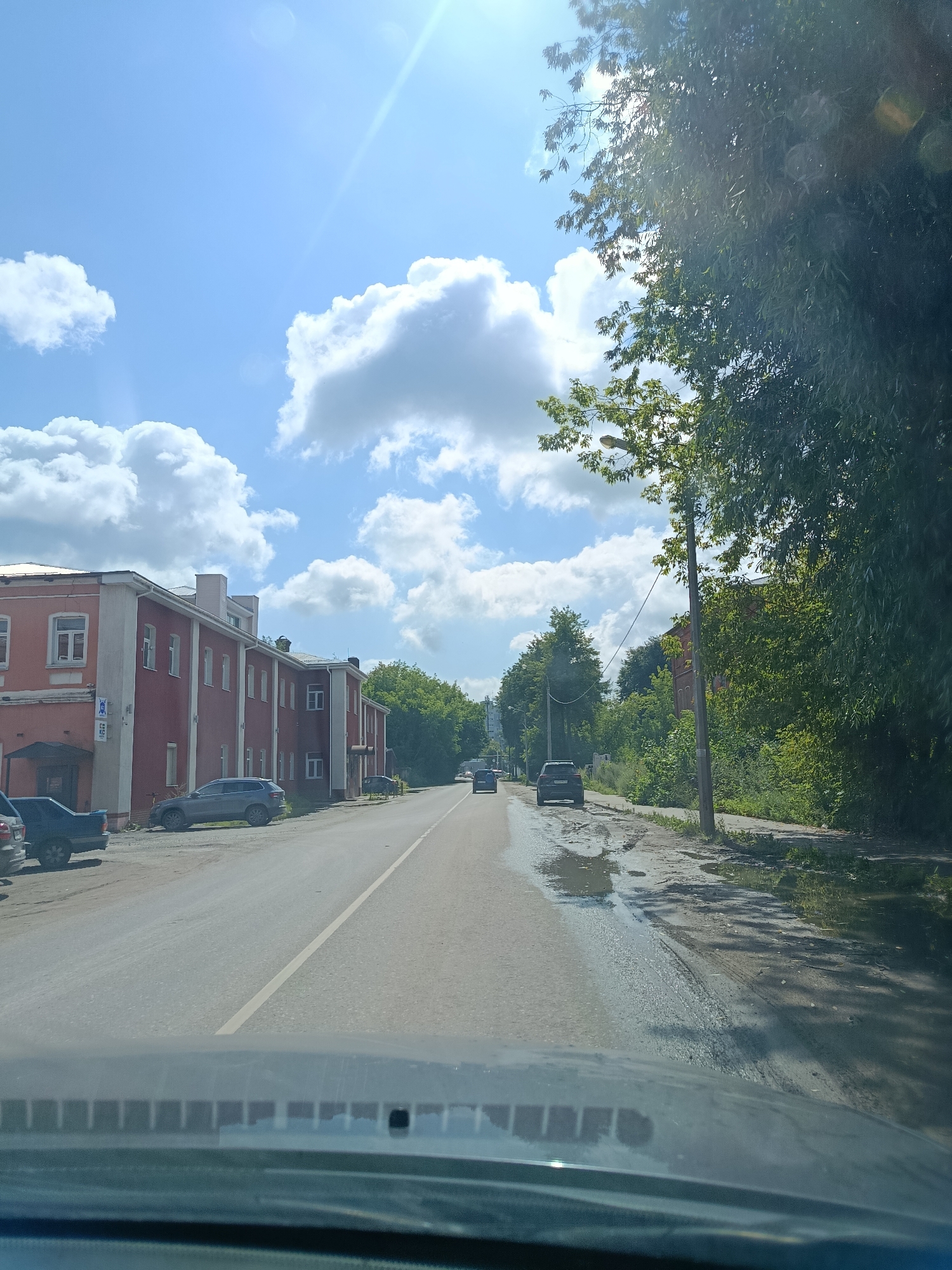 The image depicts a street scene in the city of Orekhovo-Zuyevo, Russia, on a sunny day with scattered clouds. The viewpoint is from inside a vehicle looking forward. On the left side of the street, a long, red, multi-story building with white trim and multiple windows is visible. Several cars are parked along the building. The road ahead is paved and appears to be in fair condition, with some puddles and rough patches on the right side. Green trees line the right side of the road, and some appear to be on the left side further down the street. A wooden utility pole with wires runs along the right side of the road. Several cars are driving on the road, and a few are parked. The sky is blue with fluffy white clouds, and the sun is shining brightly, creating lens flares in the upper right portion of the image.