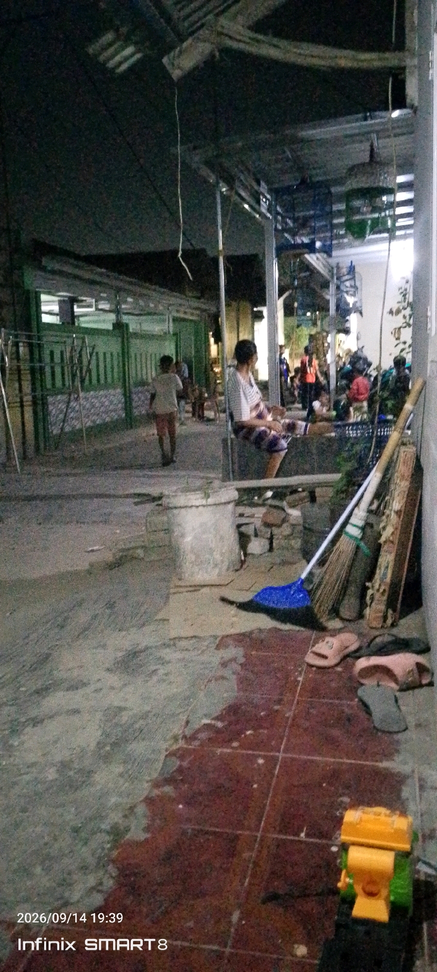 In the city of Serang, Indonesia, an outdoor scene unfolds at dusk. A walkway, paved with dark red tiles, stretches into the background, merging with a gray, uneven surface. A large concrete pot with visible vegetation sits on the walkway. To the right, a collection of brooms leans against a wall, accompanied by a blue broom lying on the ground. Several pairs of sandals are scattered near the brooms. Further into the background, a young child in a light-colored shirt and shorts walks away from the camera. To the right of the walkway, a person is seated, facing away from the viewer, wearing a striped shirt and patterned shorts. In the distance, a green fence with vertical slats encloses a building. The environment suggests a residential or communal area, possibly a back alley or courtyard, with overhead structures and lighting from within buildings contributing to the dim ambiance. A small yellow and green toy vehicle is visible on the tiled portion of the walkway in the foreground. The timestamp "2026/09/14 19:39" and "Infinix SMART 8" are present in the lower left corner.