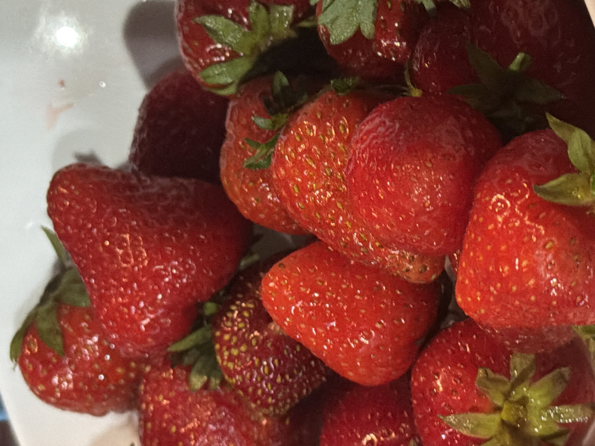 This is a close-up, overhead shot of a bowl of fresh strawberries. The strawberries are a vibrant red, with visible seeds and green stems. The lighting creates highlights and shadows, emphasizing the plumpness and texture of the fruit. There are no people or discernible environmental features beyond the immediate context of the strawberries in a bowl. The image does not provide any information that would allow for identification of a specific location in Sochi, Russia.