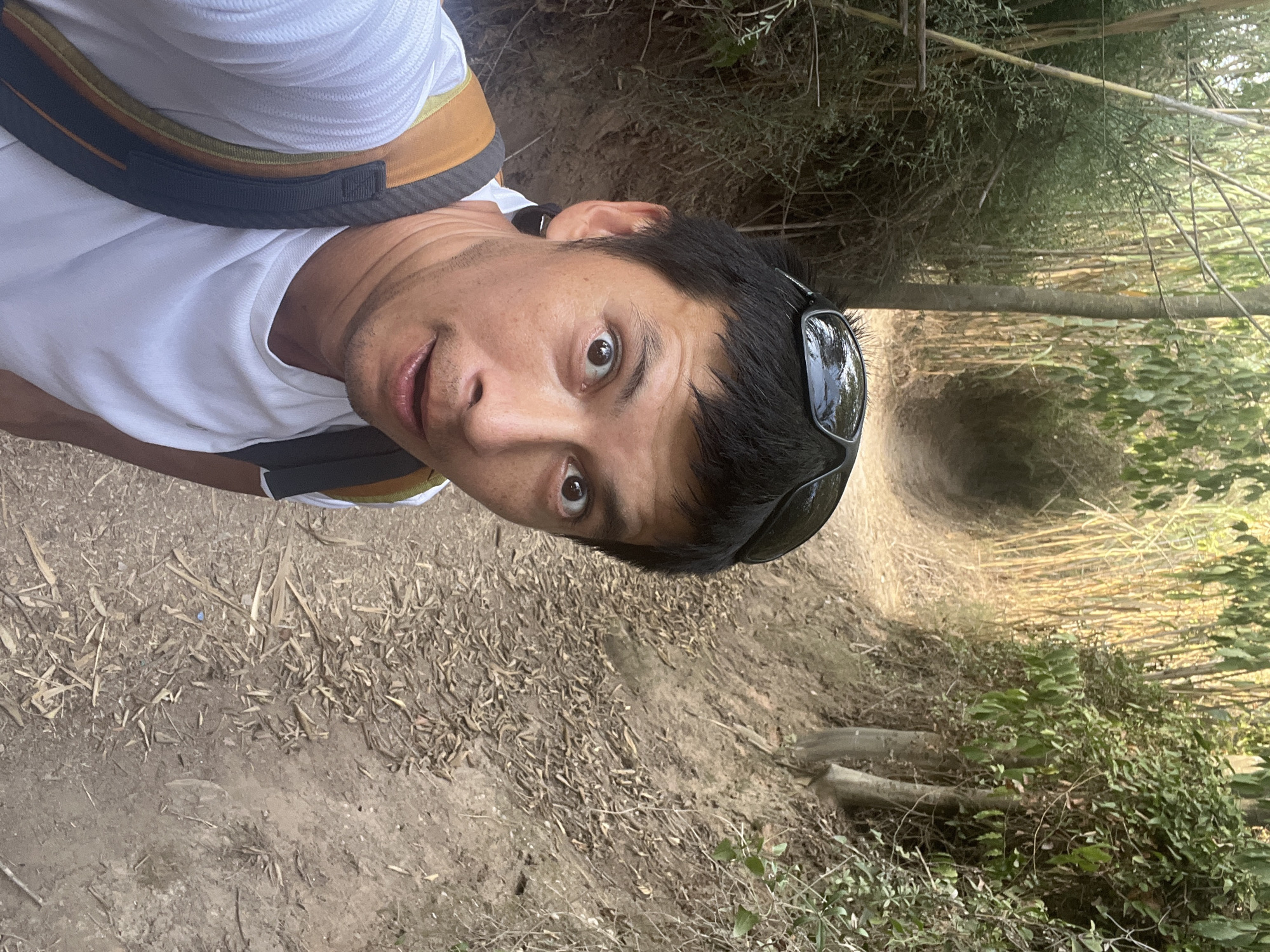 A man is taking a selfie while walking on a dirt path in a natural setting. He is wearing a white t-shirt and a backpack with orange and gray straps. Sunglasses are perched on his head. The path is flanked by trees and dense vegetation. The ground is covered with dry leaves and dirt. The background shows a narrow, winding path disappearing into the greenery. The image appears to have been taken during the day, with sunlight filtering through the trees.
