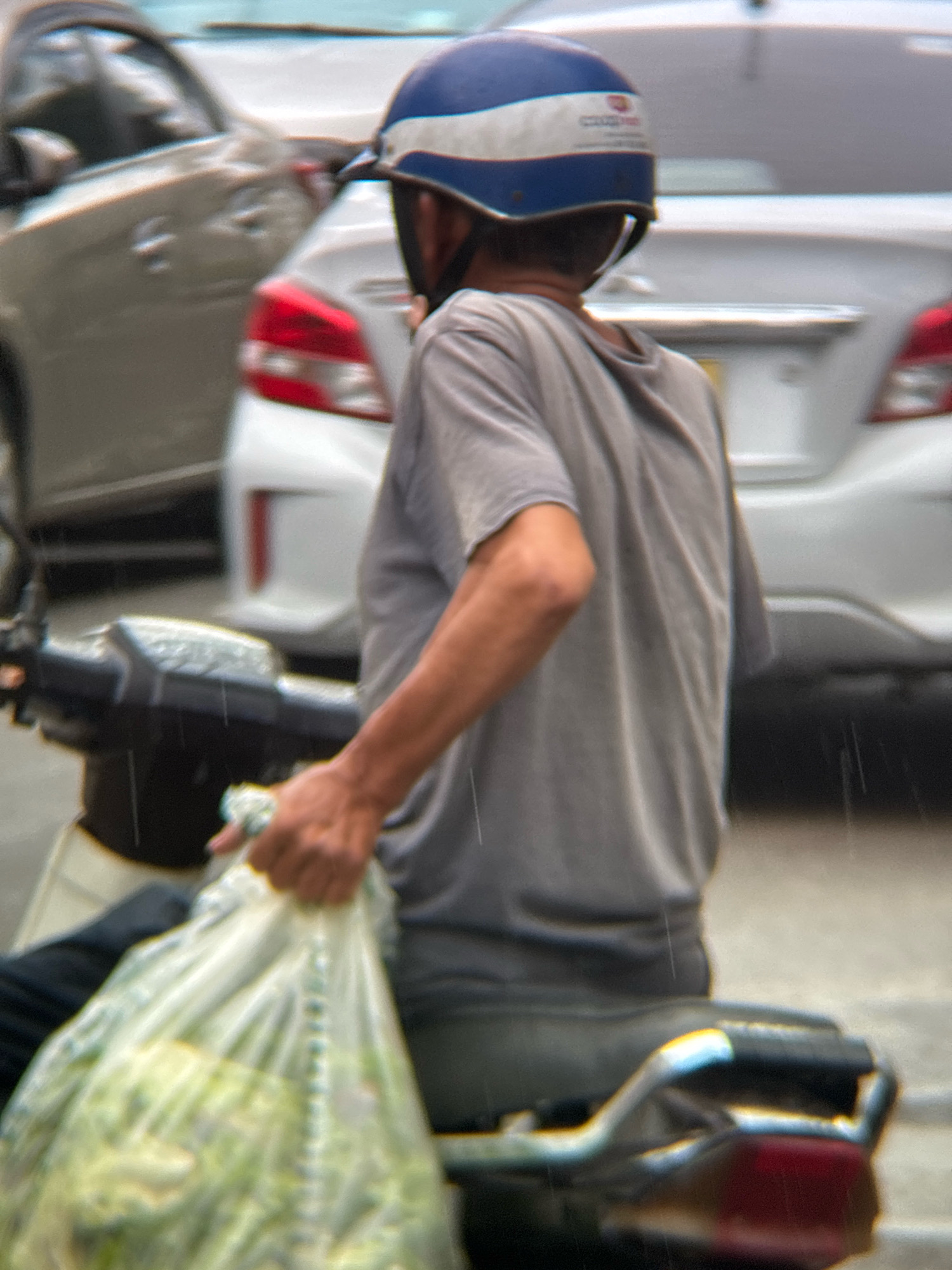 An overcast day in Nha Trang, Vietnam, with light rain falling, is captured in this close-up image of a person on a motorcycle. The individual, wearing a blue and white helmet, is dressed in a grey t-shirt and dark pants. They are holding a large, transparent plastic bag filled with what appears to be green produce. The motorcycle's handlebars and part of the seat are visible. In the background, other vehicles, including a light-colored car, are slightly blurred due to motion and the rain. The overall scene depicts a moment of everyday life in the city, likely during a commute or a trip to the market.
