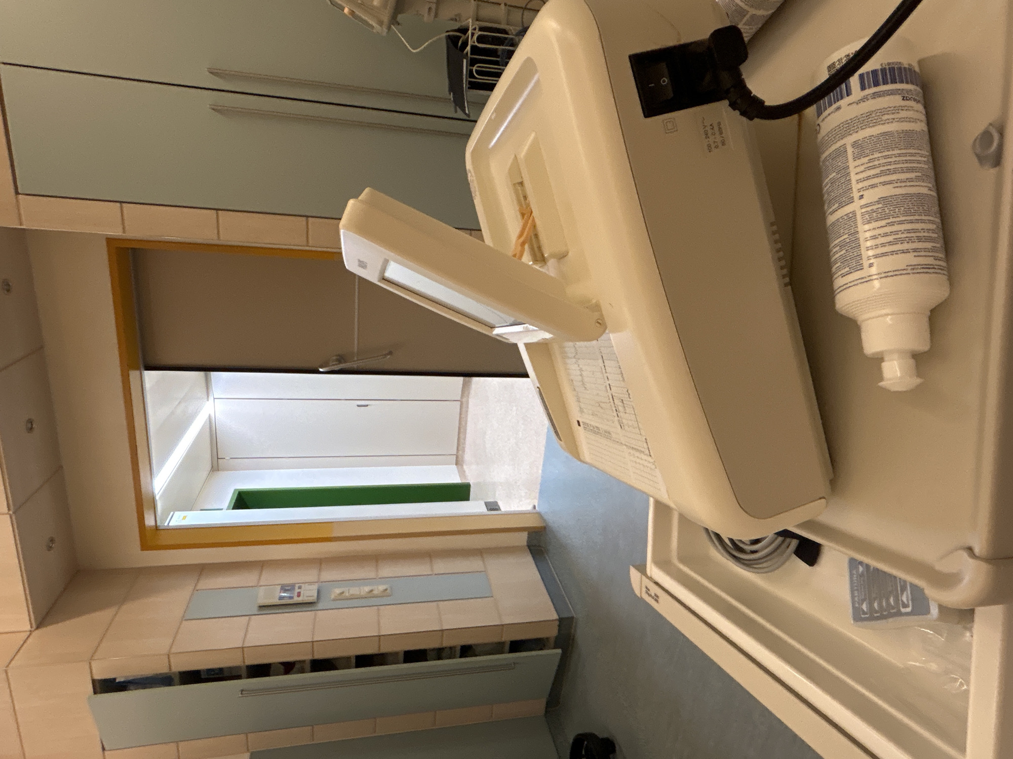 The image shows a hallway with a door in the background leading to a room. In the foreground, there is a medical device on a tray. The device is white and has a screen that is tilted upwards. A bottle of liquid is lying on the tray next to the device. The hallway walls are tiled, and there are cabinets on the left side of the image. The floor is a light gray color. The lighting is overhead and appears to be fluorescent. The scene is sterile and clinical, suggesting a hospital or medical facility. There are no people visible in the image. It is not possible to determine the specific location in Gummersbach, Germany based solely on the provided image.