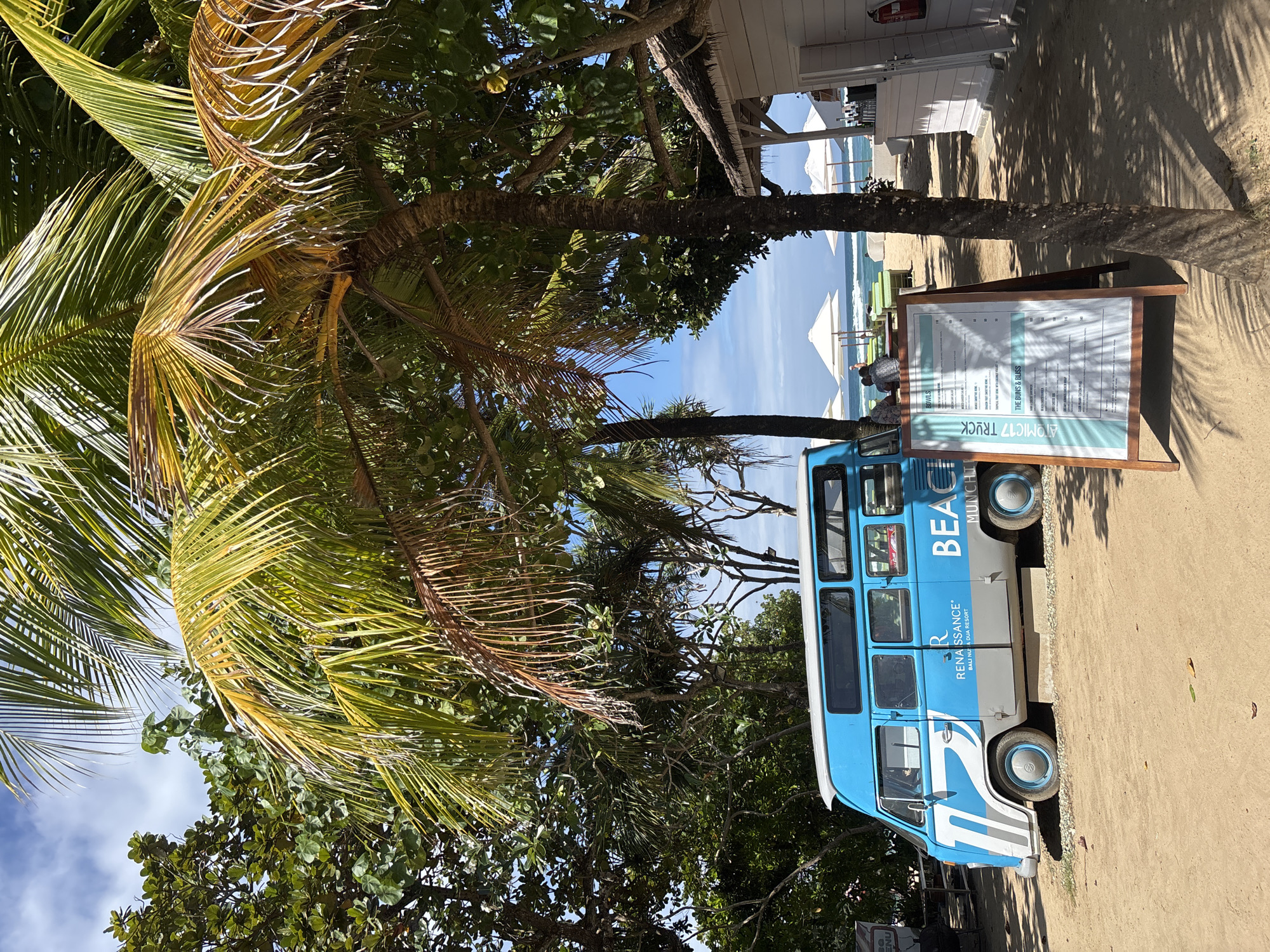 This is a sideways, eye-level shot of a blue and white shuttle bus parked on a sandy beach, with palm trees and lush green foliage in the foreground. The bus has "BEACH MUNCHIES" and "R RENAISSANCE BALI NUSA DUA RESORT" written on its side. A wooden A-frame sign stands next to the bus, displaying a menu with sections like "BOWLS" and "THE BUNS & BLISS". In the background, white beach umbrellas are set up, and the ocean with gentle waves is visible under a clear blue sky. The lighting suggests it is daytime, likely mid-morning or afternoon, with sunlight casting shadows from the trees onto the sand. The scene conveys a relaxed, tropical vacation atmosphere. The visible text on the sign is "ATOMIC17 TRUCK" and parts of the menu items.