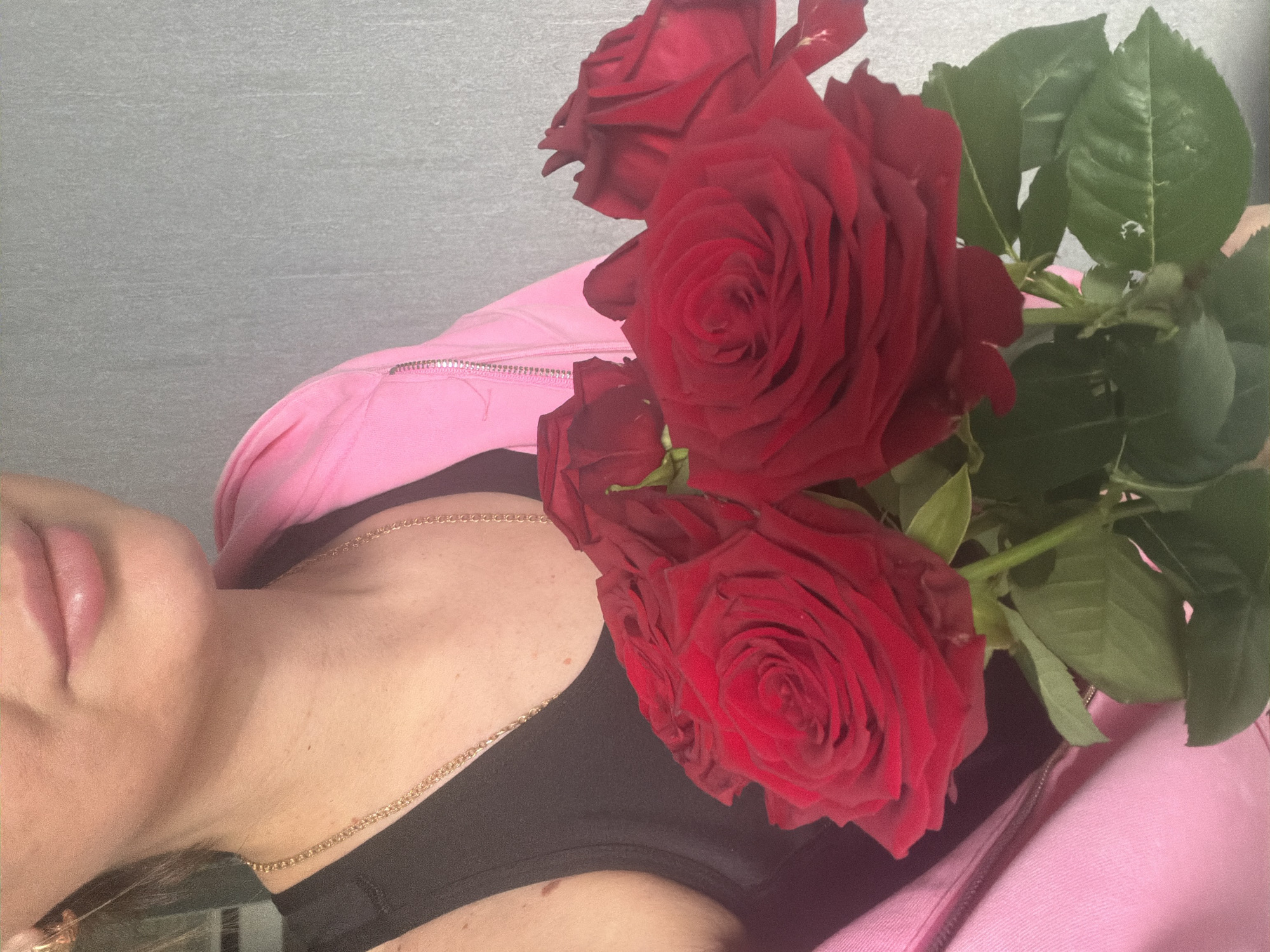 The image shows a woman's face, neck, and chest with a bouquet of red roses. She is wearing a black top and a pink jacket. A gold chain necklace is visible on her neck. The background appears to be a textured gray surface. The location cannot be determined to be Rostov-on-Don, Russia from the image alone.