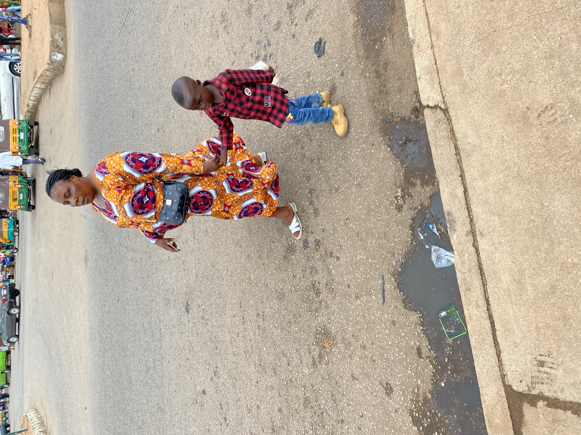 The image, taken from above, depicts a woman and a young boy walking along a street in Bukuru, Nigeria. The woman is wearing a long, patterned orange and white dress with blue accents. A black purse hangs on her side. She is wearing white sandals and holding a mobile phone. The boy is wearing a red and black checkered shirt, blue jeans, and tan-colored shoes. They are holding hands.
The street is paved, with a concrete sidewalk bordering it. There is some litter and a small puddle near the curb. In the background, there are several auto rickshaws and a white car are parked on the opposite side of the street.