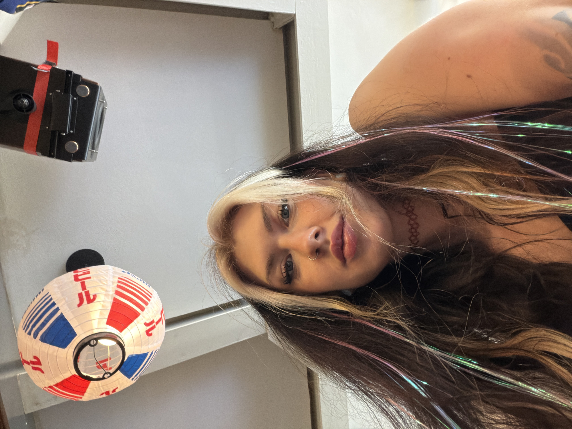 A low-angle shot reveals a woman with blonde highlights, dark brown hair and colorful strands of tinsel, with her right shoulder visible. She has a nose ring and a choker. In the background, there is a white paper lantern with Japanese writing and red and blue patterns, and a black object with red tape. The lantern is hanging from the ceiling. A gray wall and trim are also visible in the background. There is no indication of this image being taken in Belgrade, Serbia.