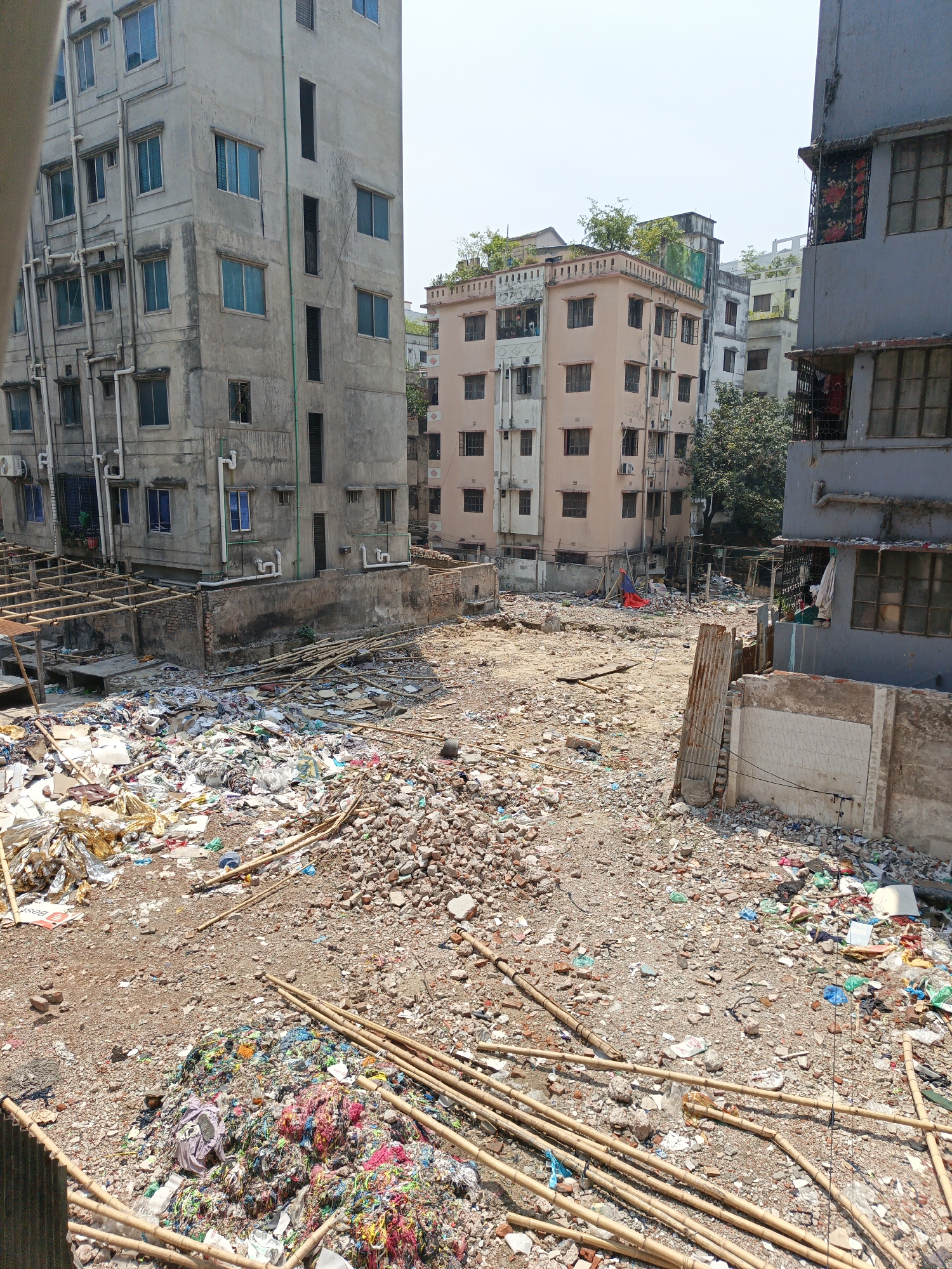 The image depicts a vacant lot filled with debris and garbage in Dhaka, Bangladesh. The foreground shows a mix of household trash, construction materials, and organic waste. There are bamboo poles scattered across the ground and piles of textile waste. In the background, there are several multi-story buildings with faded paint and exposed piping. Some buildings have greenery on their rooftops. The overall environment suggests a densely populated urban area with inadequate waste management infrastructure. There are no people visible in the scene.