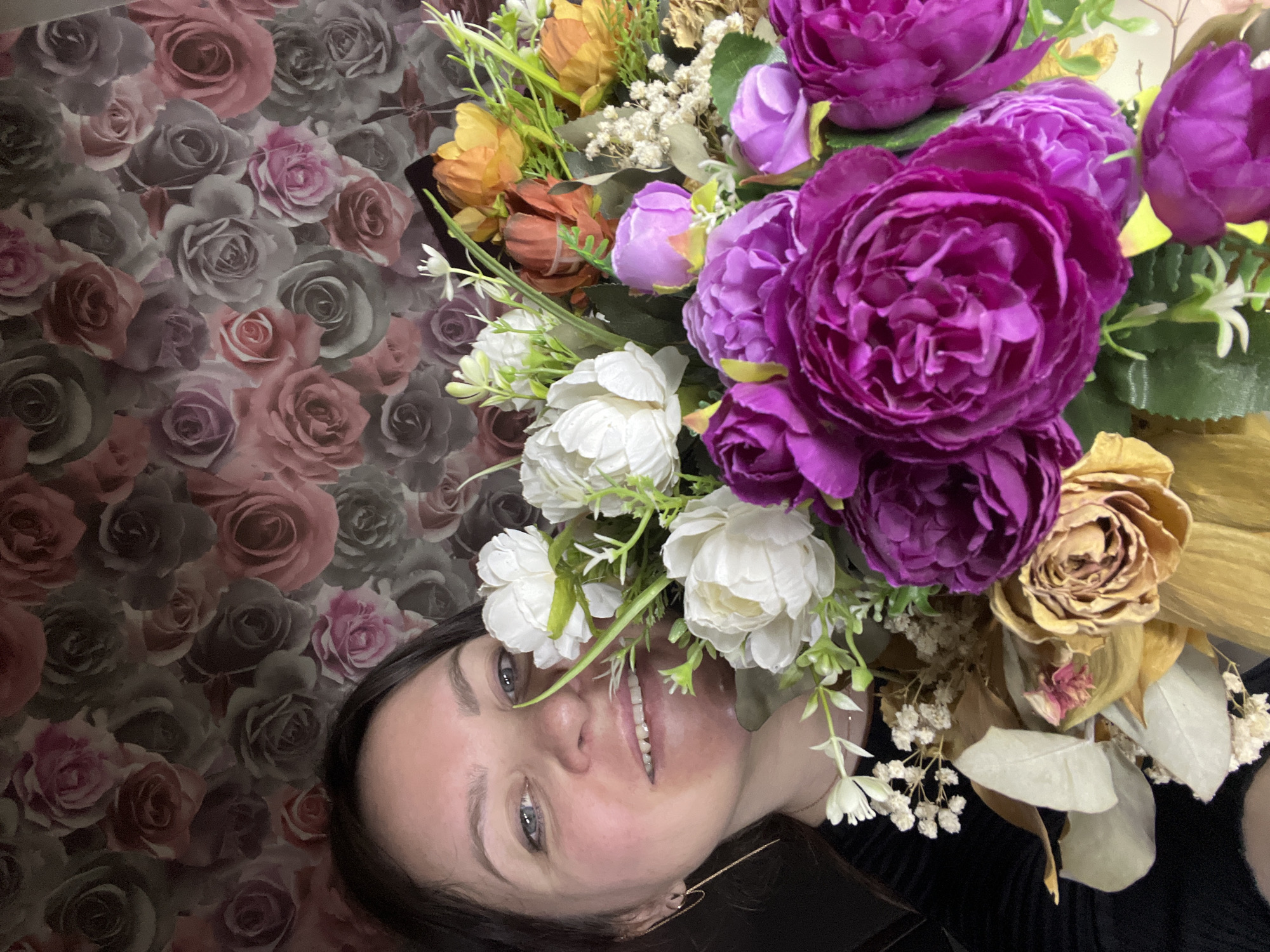 A woman smiles up at the camera while surrounded by a bouquet of flowers, with a wallpaper patterned with roses as a backdrop. The woman has dark hair, fair skin, and is wearing a black top. The bouquet contains various colors of flowers, including purple, white, orange, and yellow. The scene is likely indoors. There is no information available to determine that the scene is in Noviy, Russia.
