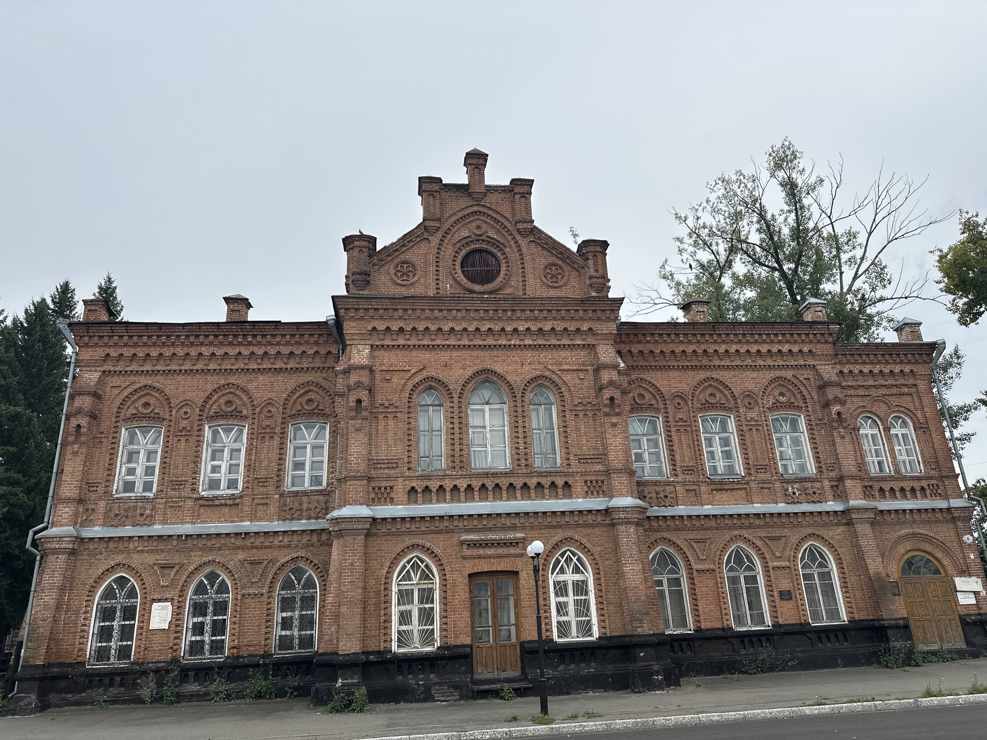 The image shows a detailed, front-facing view of the historic Biysk City Drama Theater, located in Biysk, Russia. The building is constructed of red brick with ornate architectural details, including arched windows, decorative brickwork, and a central gable. The sky is overcast and gray. Trees are visible to the left and right of the building, and a street runs in front of it. There are no people visible in the image.