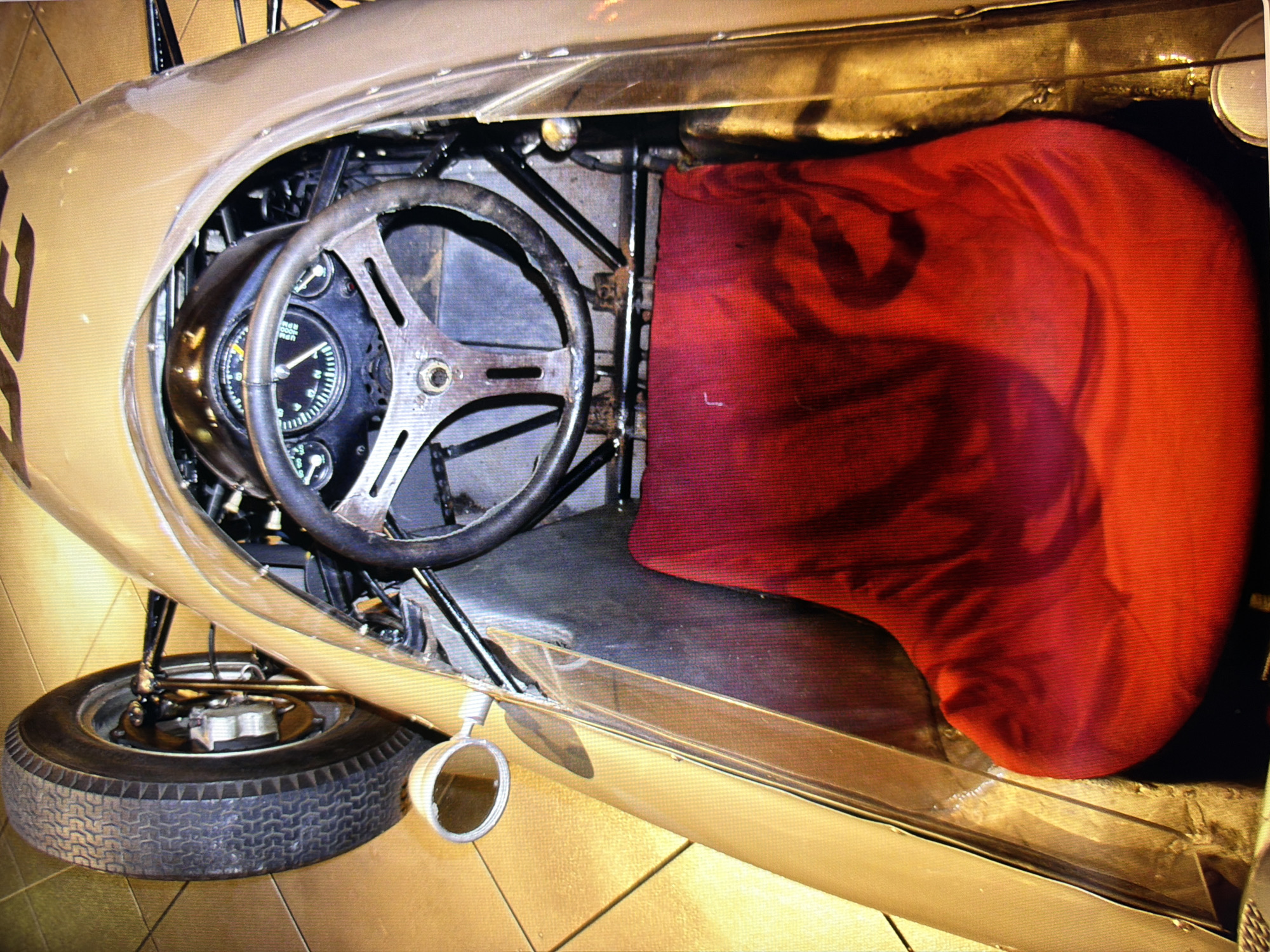 The image shows a top-down view of the interior of a vintage racing car, likely on display in a museum or exhibition. The car is predominantly gold or champagne colored. In the foreground, a plush red fabric covers what appears to be a racing seat, its contours visible beneath the material. To the left, a section of the car's gold exterior is visible, meeting the black tire of the front wheel, which is partially obscured. The tire has a classic tread pattern. Directly behind the steering wheel, a dashboard with gauges is visible, the needles indicating specific readings. The steering wheel itself is black and appears worn, with a metal central hub. The overall impression is one of vintage motorsport. The location is stated to be in the city of Kyiv, Ukraine.