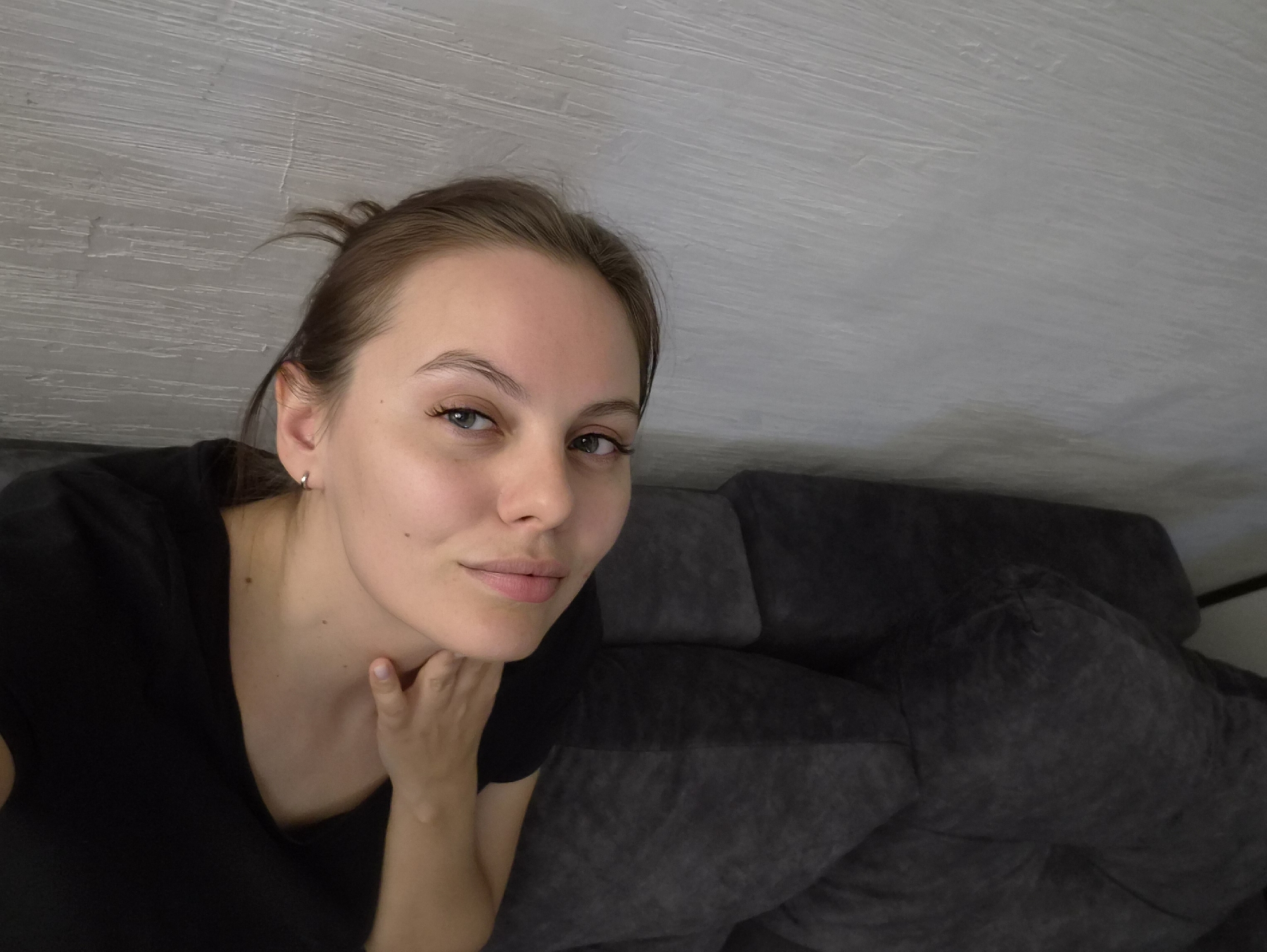 This is an indoor selfie of a young woman taken from a slightly high angle. The primary subject is a young woman with light brown hair, pulled back but with some wisps framing her face, and light-colored eyes. She is looking directly into the camera with a neutral to slightly pleasant expression, exhibiting a subtle hint of a smile. She is wearing a black t-shirt and has a small silver hoop earring in her visible left ear. Her right hand is gently resting under her chin/jawline.
In the background, to the right of the woman, is a dark grey, upholstered sofa or couch. Above and behind her, a light grey wall or ceiling with a visible textured finish is present. The lighting suggests it is daytime. The overall setting appears to be a casual indoor environment, consistent with a home in Chaikovsky, Russia. There is no visible text in the image.