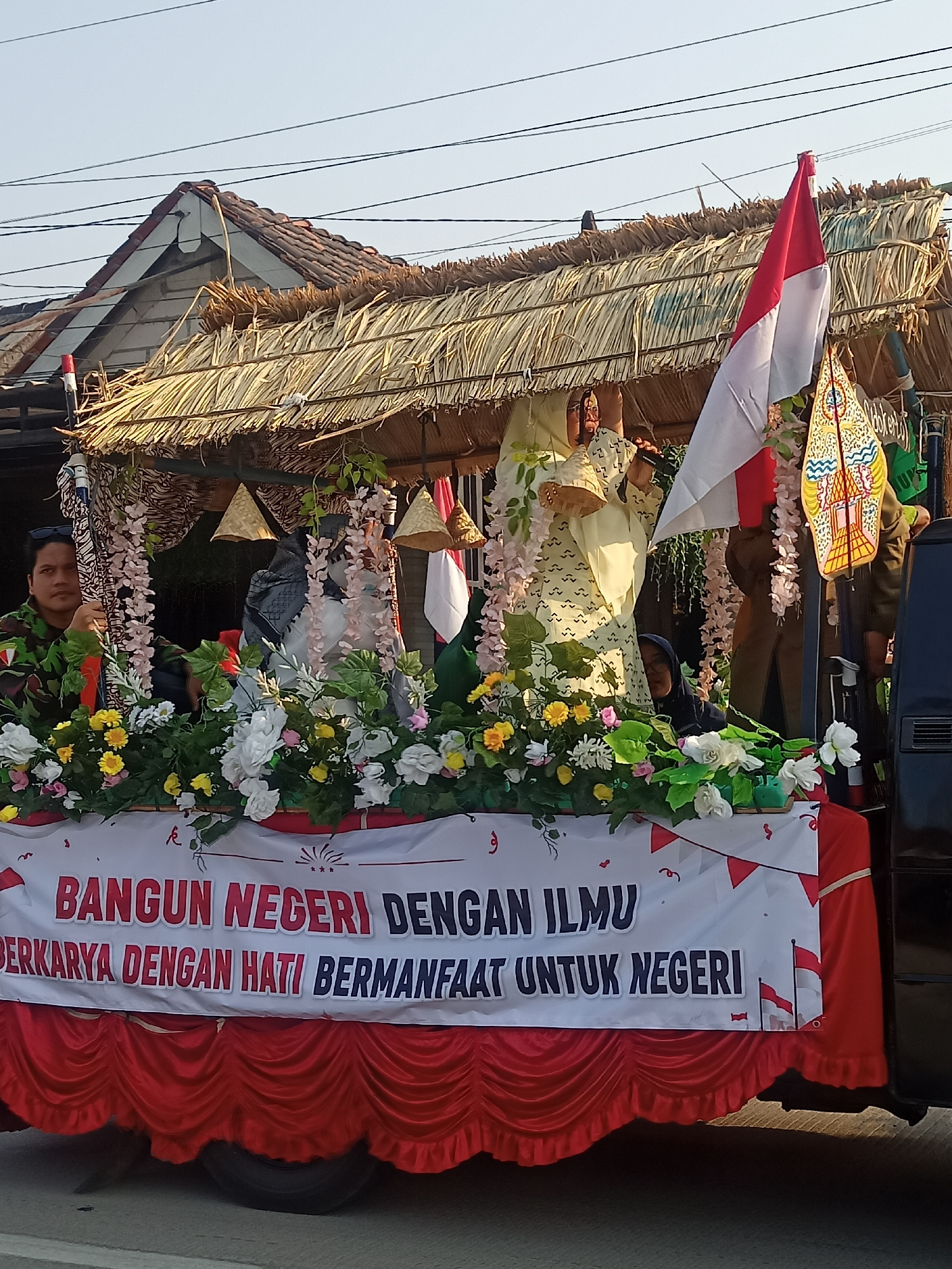 The image depicts a decorated float in the city of Ngrompongan, Indonesia, likely for a parade or celebration. The float features a thatched-roof structure, reminiscent of a traditional house or stall, adorned with various decorations. Prominently displayed is the Indonesian flag, a red and white bicolor. Several people are visible on the float, including a man in a camouflage uniform on the left, a woman in a patterned yellow and white outfit in the center holding a basket, and another person wearing a dark head covering behind the flowers. The front of the float is draped with a large banner that reads "BANGUN NEGERI DENGAN ILMU BERKARYA DENGAN HATI BERMANFAAT UNTUK NEGERI," which translates to "BUILD THE NATION WITH KNOWLEDGE, WORK WITH HEART, BENEFIT THE NATION." The float is decorated with an abundance of artificial flowers in white, yellow, and pink, as well as green foliage. Small, cone-shaped straw decorations hang from the structure. In the background, a building with a tiled roof is visible, along with overhead power lines against a clear sky. The float is positioned on a street, with the lower portion of a dark-colored vehicle visible on the right side of the frame.