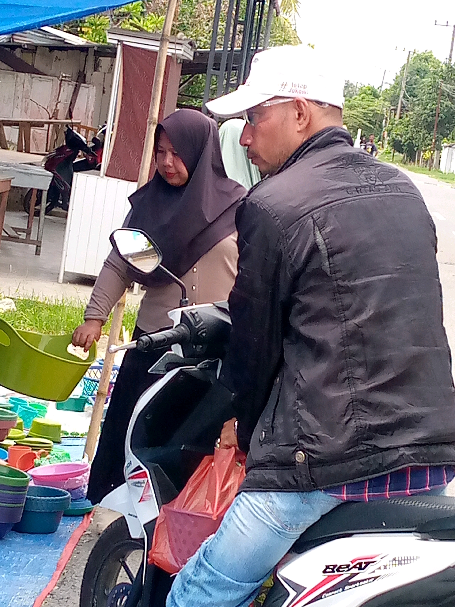 In Keudegeudong, Indonesia, a woman wearing a dark hijab and a light brown shirt is handing a white cloth to a man. The man, wearing a white baseball cap with text and glasses, is seated on a motorcycle and has his back to the camera. He is wearing a black jacket over a plaid shirt and blue jeans. The motorcycle is parked on a blue tarp, which is also covered with various colorful plastic basins and containers. In the background, there is a building with wooden elements, bamboo poles, and lush green vegetation. The scene appears to be an outdoor market.