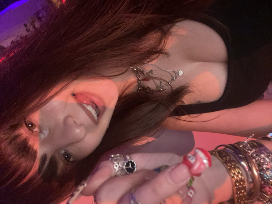 A woman with dark, fringed hair and light skin is angled toward the viewer. She is wearing a low-cut black top and several layered necklaces. Her tongue is slightly visible as she smiles. Her hand is adorned with multiple rings and a stack of gold bracelets. Her nails are painted in a light color. The background lighting is a mix of pink and purple tones.
