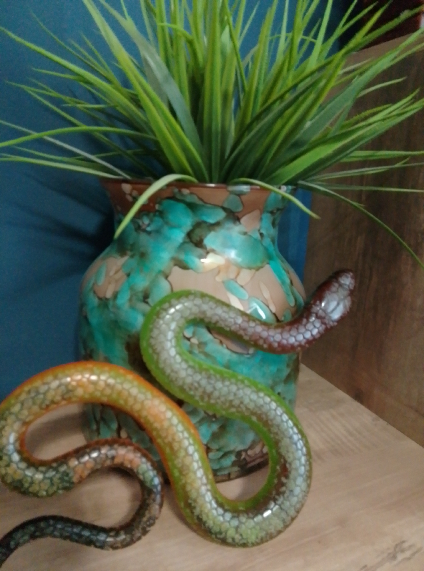 The image shows a decorative serpentine sculpture coiled around a vase. The vase is ornate with a speckled turquoise and brown glaze, and it contains a large, spiky green plant. The serpentine sculpture is coiled in several loops, with its head near the top of the vase, and its body extending down and to the left. The sculpture is colored in shades of green, orange, and brown, and has a textured, scaled appearance. The background is a deep blue wall on the left and a wooden surface on the right. The scene appears to be an indoor still life arrangement. The location in Wedel, Germany is not discernible from the image.