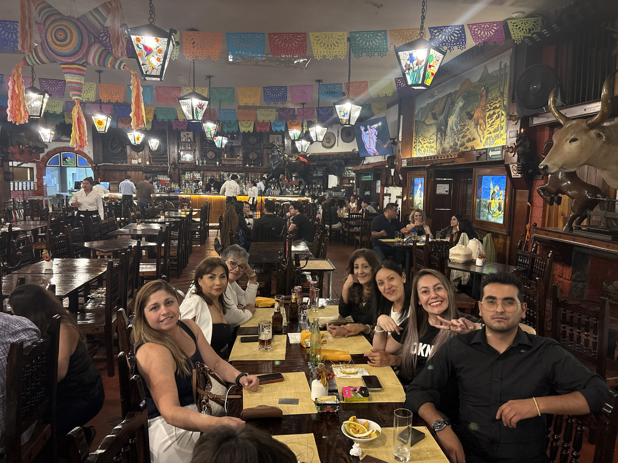 The image shows a group of people seated around a wooden table in a restaurant, presumably in Mexico City, Mexico, given the festive decorations overhead, including paper cut-out banners and lanterns. A piñata hangs from the ceiling. Most of the people at the table are looking towards the camera, with some smiling. Drinks and small plates of food are visible on the table. In the background, there is a bar area with servers and other customers. The interior of the restaurant has rustic, wooden decor, including a mural on the wall and animal head decor.