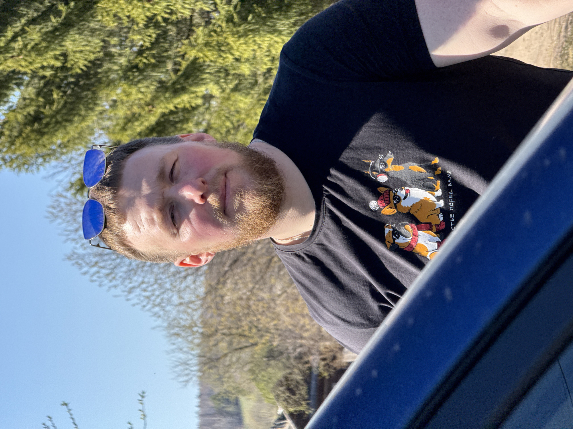A man with a beard and sunglasses perched on his head is pictured outdoors. He is wearing a dark t-shirt featuring cartoon dog designs. The scene is partially obscured by the blue frame of what appears to be a car window or vehicle interior. In the background, trees are visible under a clear, blue sky. The specific location is not definitively identifiable as Nizhnyaya Chermoda, Russia from the information provided within the image.