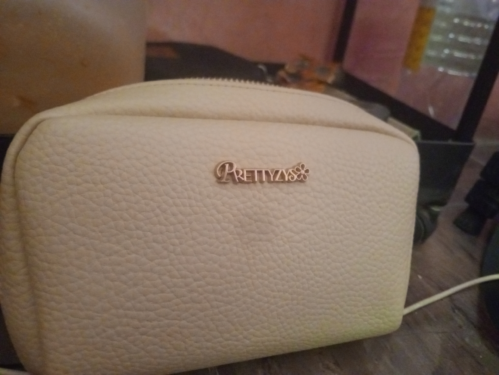 A beige, textured cosmetic bag sits on a dark wooden surface. A small, rose-gold colored emblem with the word "PRETTYLYS" and a flower design is attached to the front of the bag. In the background, out of focus, is a cylindrical object, possibly a vase or container, with a light brown hue. To the right of the cosmetic bag, the scene is dominated by the corner of a black object with a glass enclosure, possibly an aquarium or terrarium, containing a blurry yellow object and other indistinct items. A thin white cord extends from the bottom right of the frame. The image does not provide any information about the location in Bloro, Indonesia.