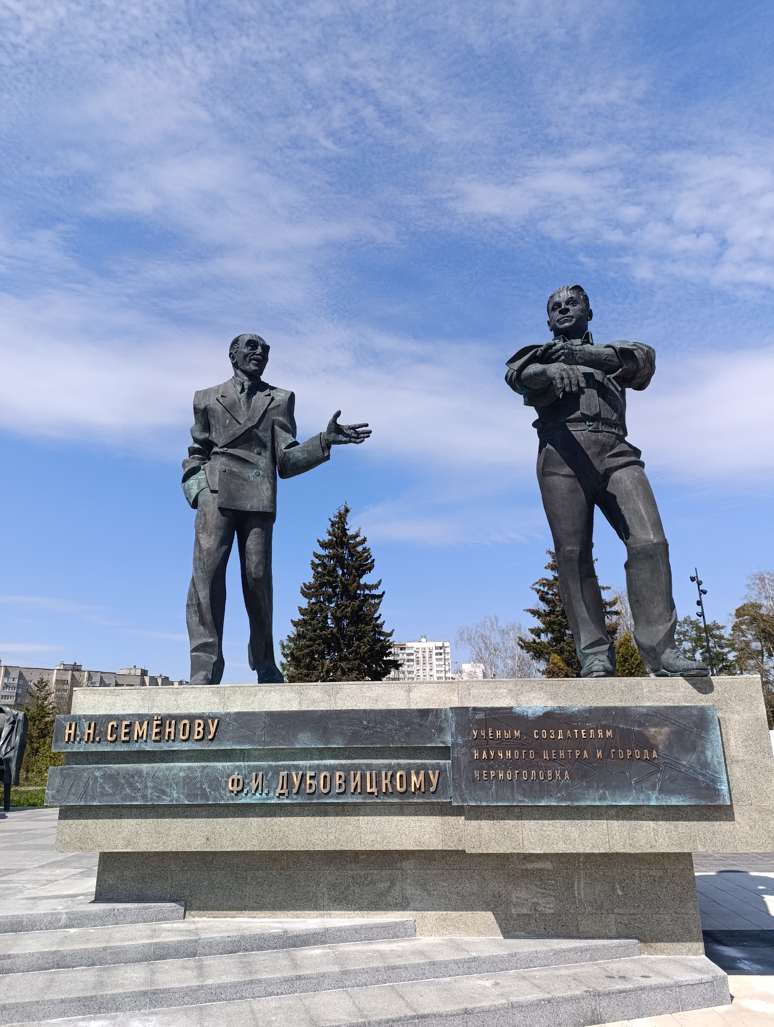 This image shows a bronze monument of two men, set against a bright blue sky with scattered clouds. The monument stands on a multi-tiered stone base with inscriptions. The man on the left wears a suit jacket, with one hand in his pocket and the other extended outwards. The man on the right wears a shirt and trousers, with his arms crossed over his chest. Behind the monument are several trees, including a prominent conifer. In the background, buildings are visible in the distance. Based on the inscriptions, the monument honors N.N. Semenov and F.I. Dubovitsky and is located in Chernogolovka, Russia.