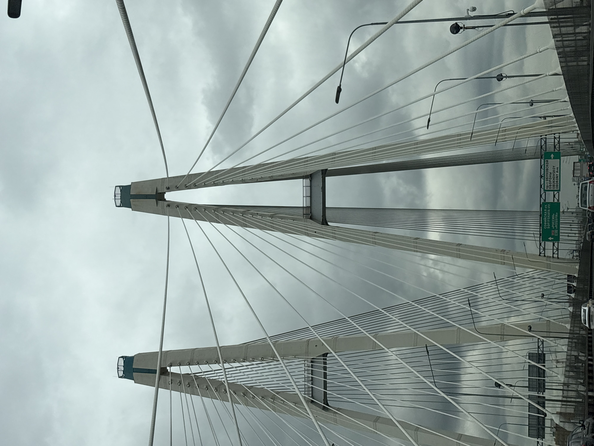The image shows a cable-stayed bridge with two towers against a cloudy sky. The towers are light gray with blue accents at the top. Numerous white cables extend from the towers to the bridge deck. Streetlights line the side of the bridge. Road signs indicate directions to Sofiyskaya Street and Obukhovsky Oborony Avenue, and the word "Rossiya", indicating that the image was most likely taken near Novosaratovka, Russia. Cars are visible on the road deck.