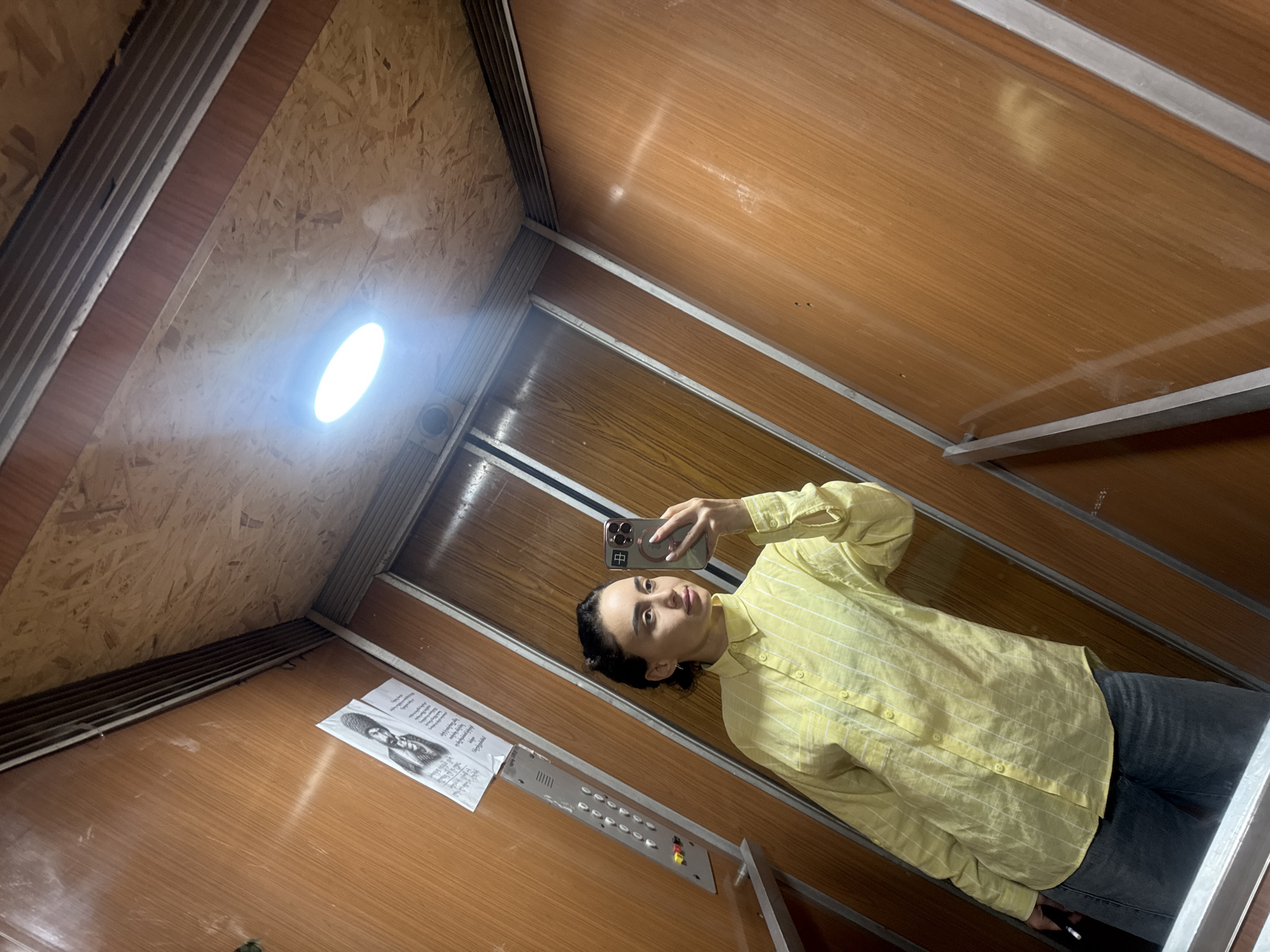 A woman is taking a selfie in what appears to be the interior of a freight elevator. The elevator is lined with wooden panels and a sheet of paper with a black and white photo is pinned to the side. The woman is wearing a yellow striped shirt and jeans, and she's holding a smartphone up to take the photo. The elevator has a light in the ceiling and a control panel on the wall. The elevator looks old and is likely found in an older building in Tbilisi, Georgia.